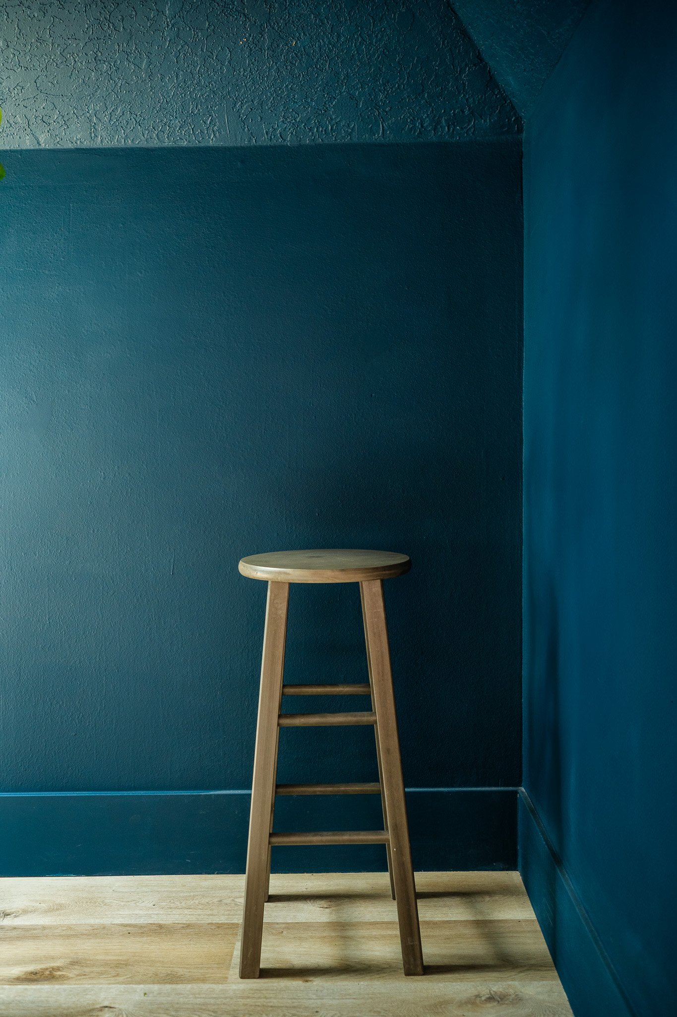 A wooden stool placed against a dark teal wall in a corner of a room with light wooden flooring.