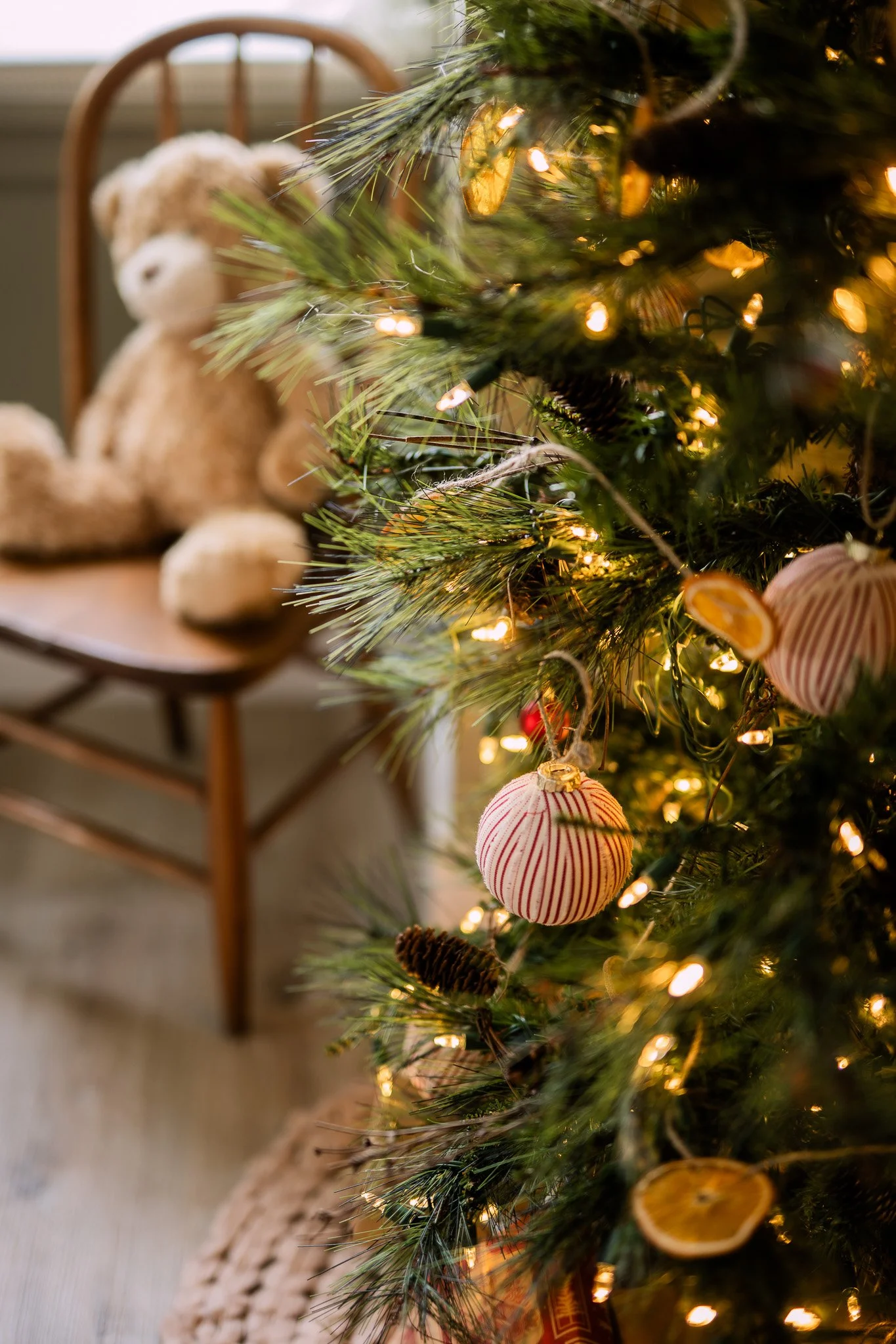 A decorated Christmas tree with lights, gingerbread ornaments, and dried citrus slices. In the background, a plush teddy bear is sitting on a wooden chair.