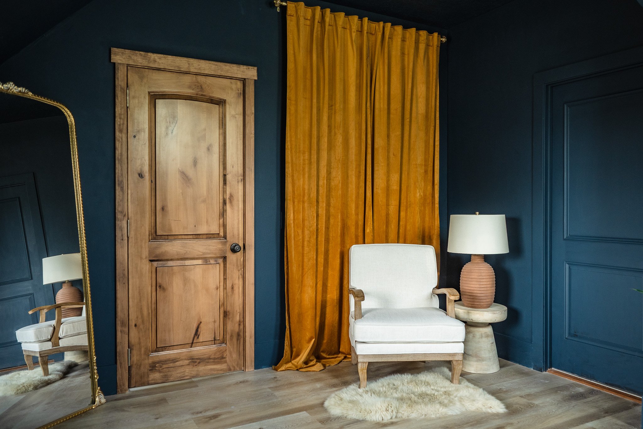 Interior of a room with dark blue walls, a wooden door, yellow curtains, a cream armchair, a table lamp on a side table, a large mirror, and a white fluffy rug on wooden floor.