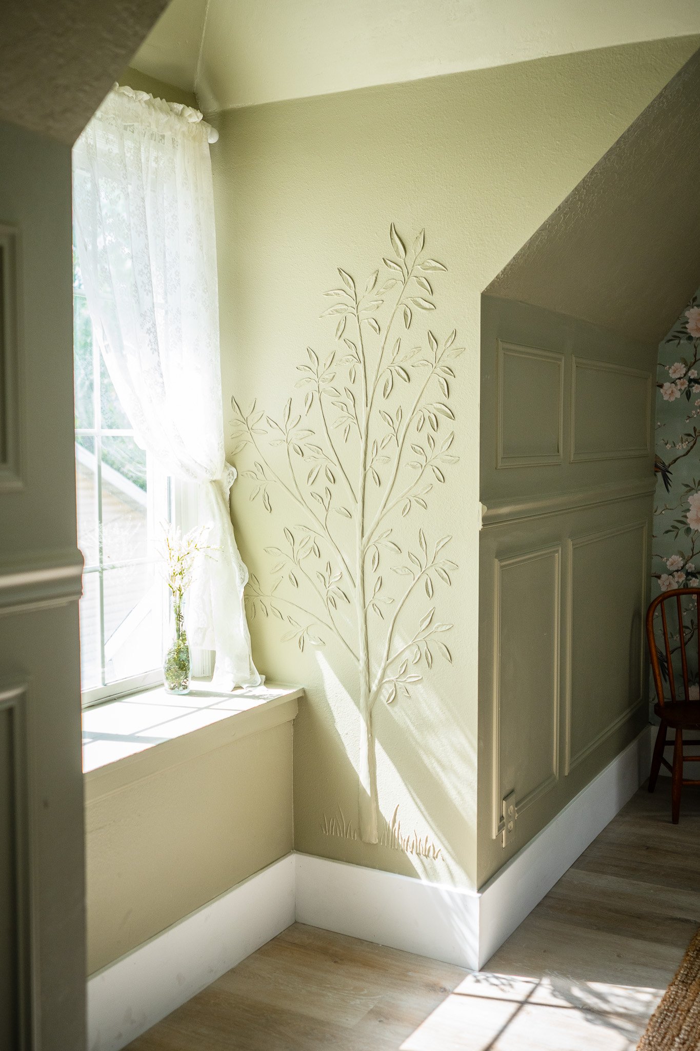 Sunlit corner of a room with a white lace curtain, a small glass vase with flowers on the windowsill, and a decorative tree shadow pattern on the wall next to a window.