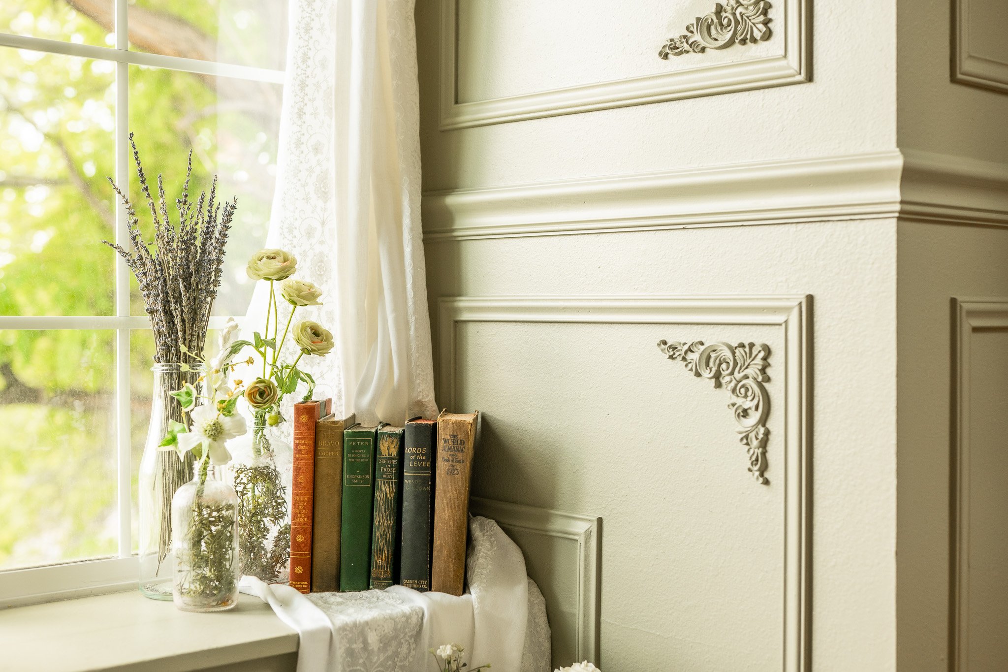 A windowsill with a glass jar of lavender, a small vase of white flowers and green foliage, a stack of vintage books, and a lace curtain. The window shows green trees outside.