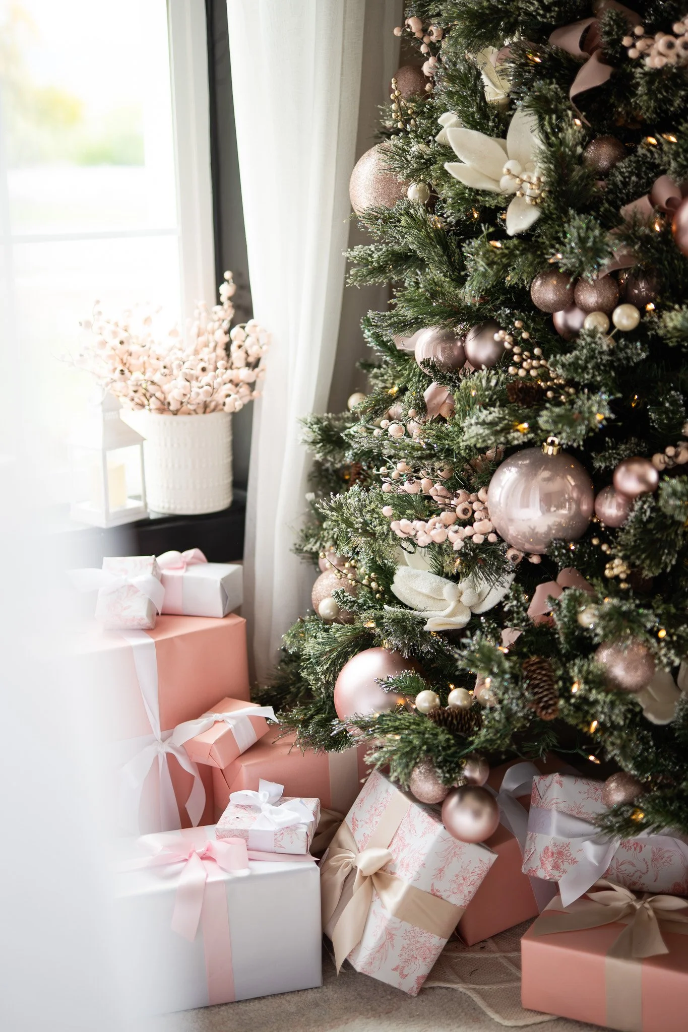 A decorated Christmas tree with pink, white, and gold ornaments. Wrapped presents in pink, white, and patterned paper with ribbons sit underneath the tree. A window with white curtains and a white vase with pink cotton branches are in the background.