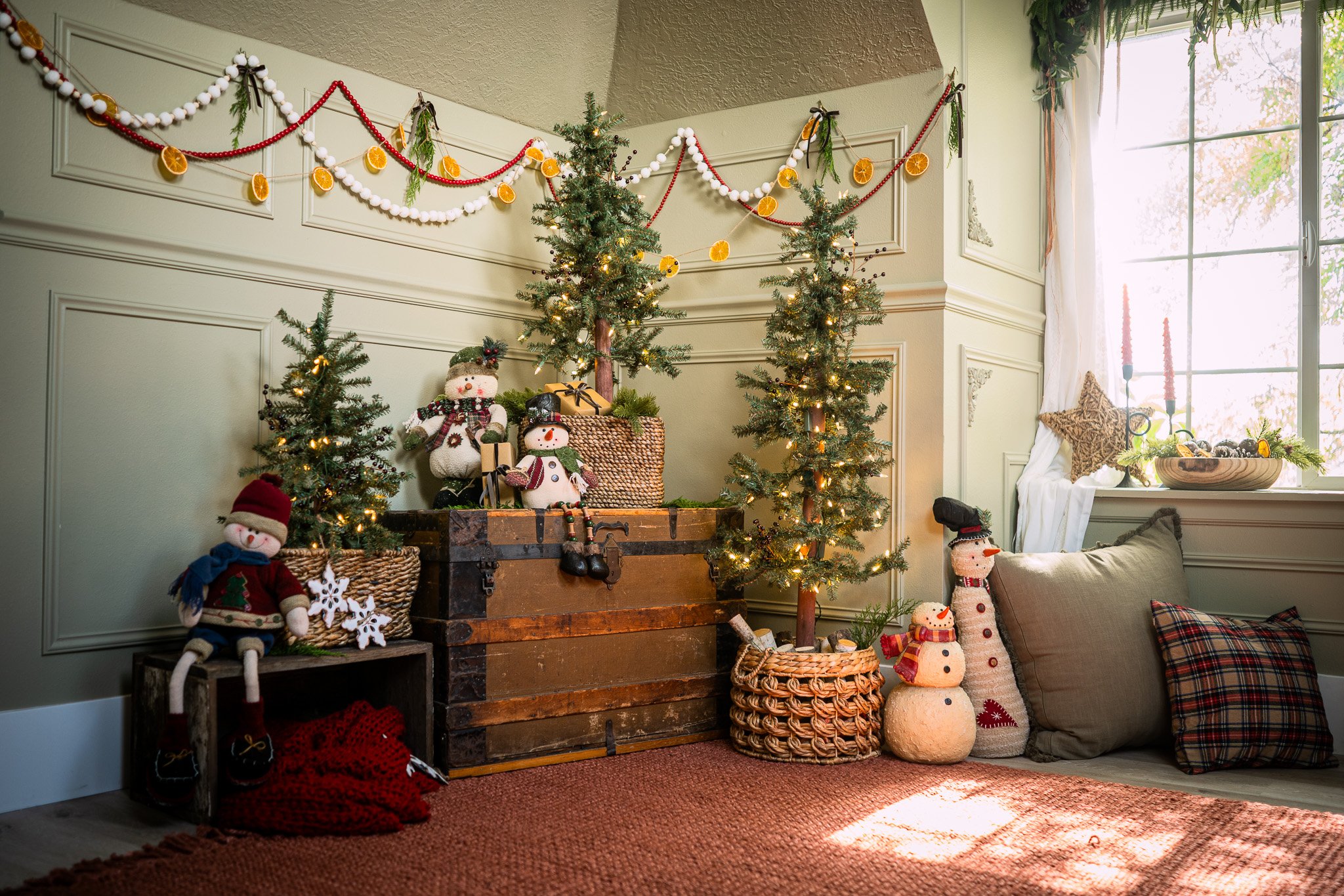Christmas decorations with small lit Christmas trees, snowman plush figures, ornaments hanging from the wall, and a window with sunlight.