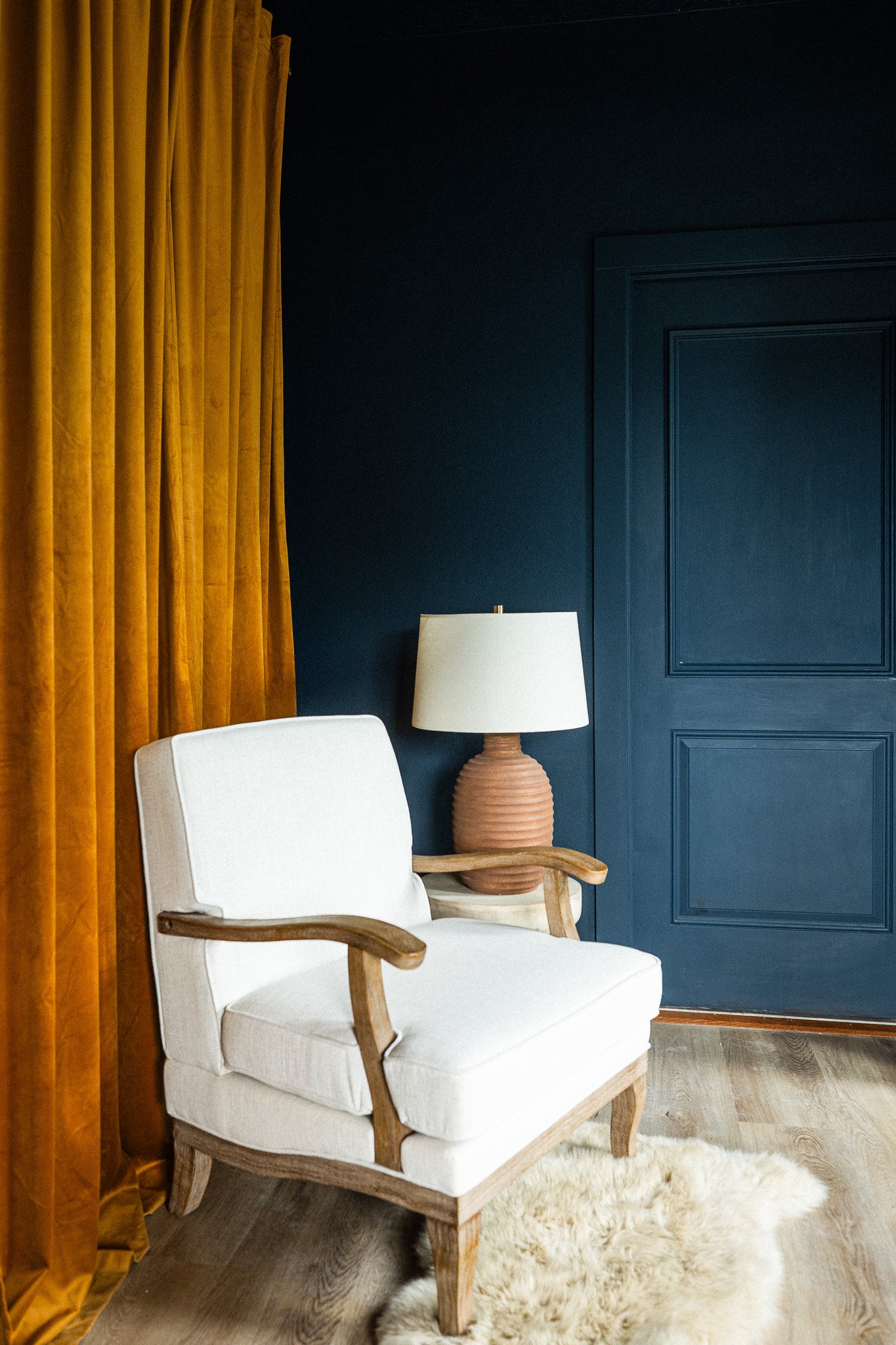A cream-colored armchair with wooden armrests and legs, positioned near a fluffy beige rug on a wooden floor, with a dark blue wall, yellow curtains, and a large beige table lamp with a ribbed terracotta base in the background.