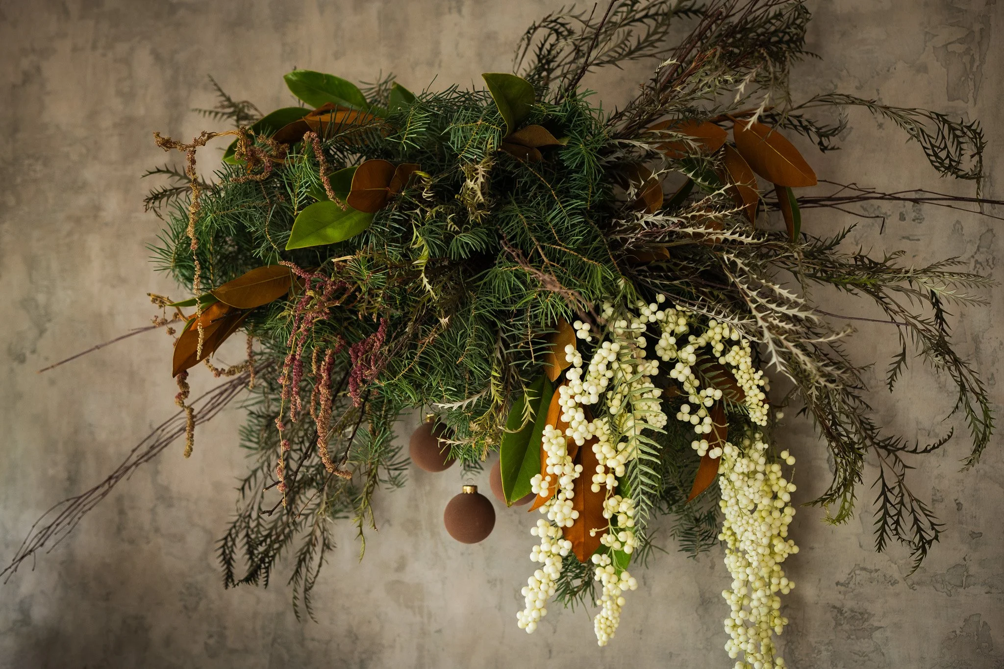 A lush holiday floral arrangement with greenery, white berries, brown leaves, and small brown ornaments hanging among the foliage.