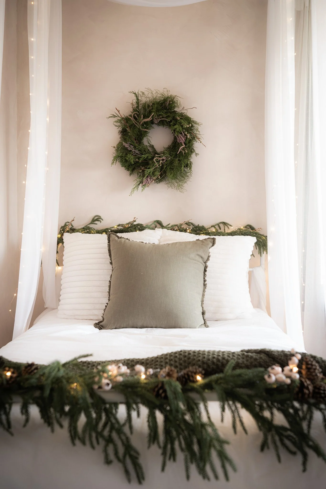 A bed decorated with evergreen garland, pillows, and candles, with a wall wreath above it, in a cozy, holiday-themed bedroom.