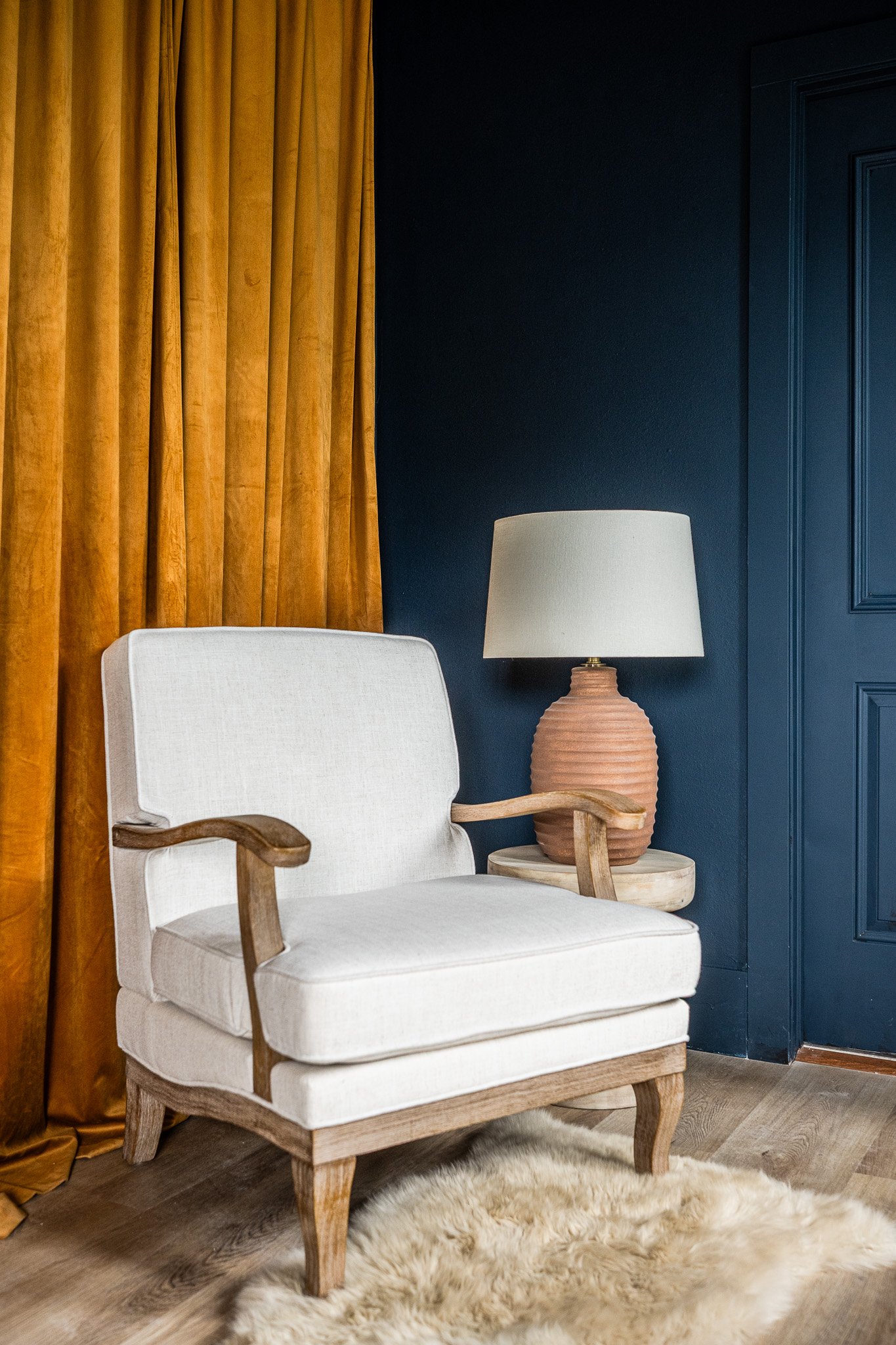 A corner of a room with a white armchair, a wooden side table, a beige table lamp, blue walls, a yellow curtain, and a fluffy rug on the wooden floor.