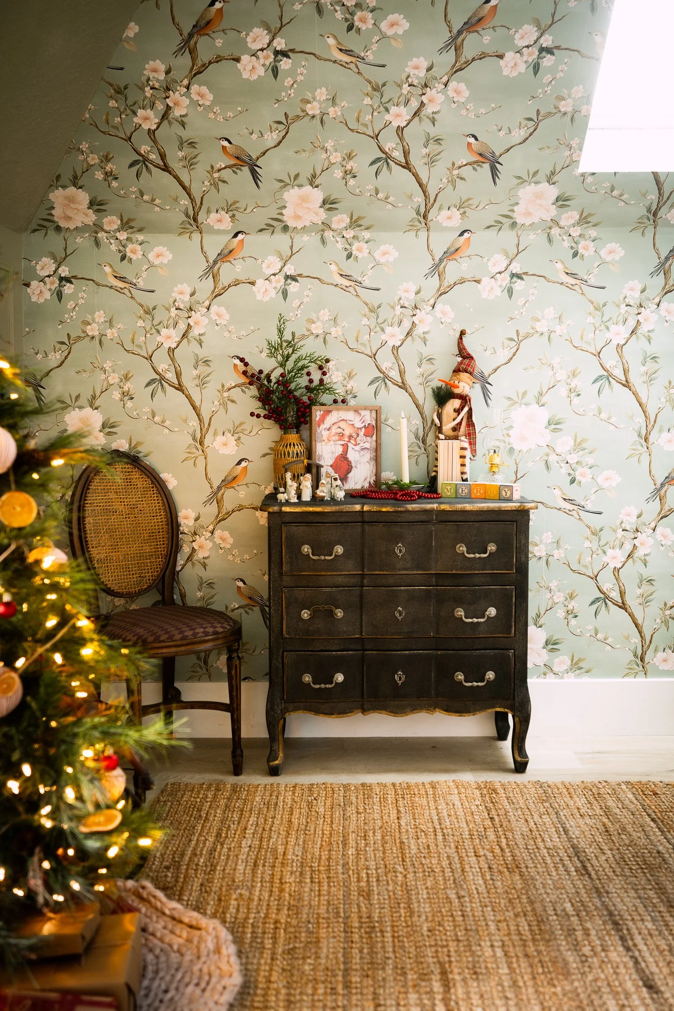 A corner of a living room decorated for Christmas with a Christmas tree on the left, a black vintage dresser facing forward, decorated with holiday ornaments, a framed Santa picture, a nutcracker, and holiday decorations on top. The room features wal