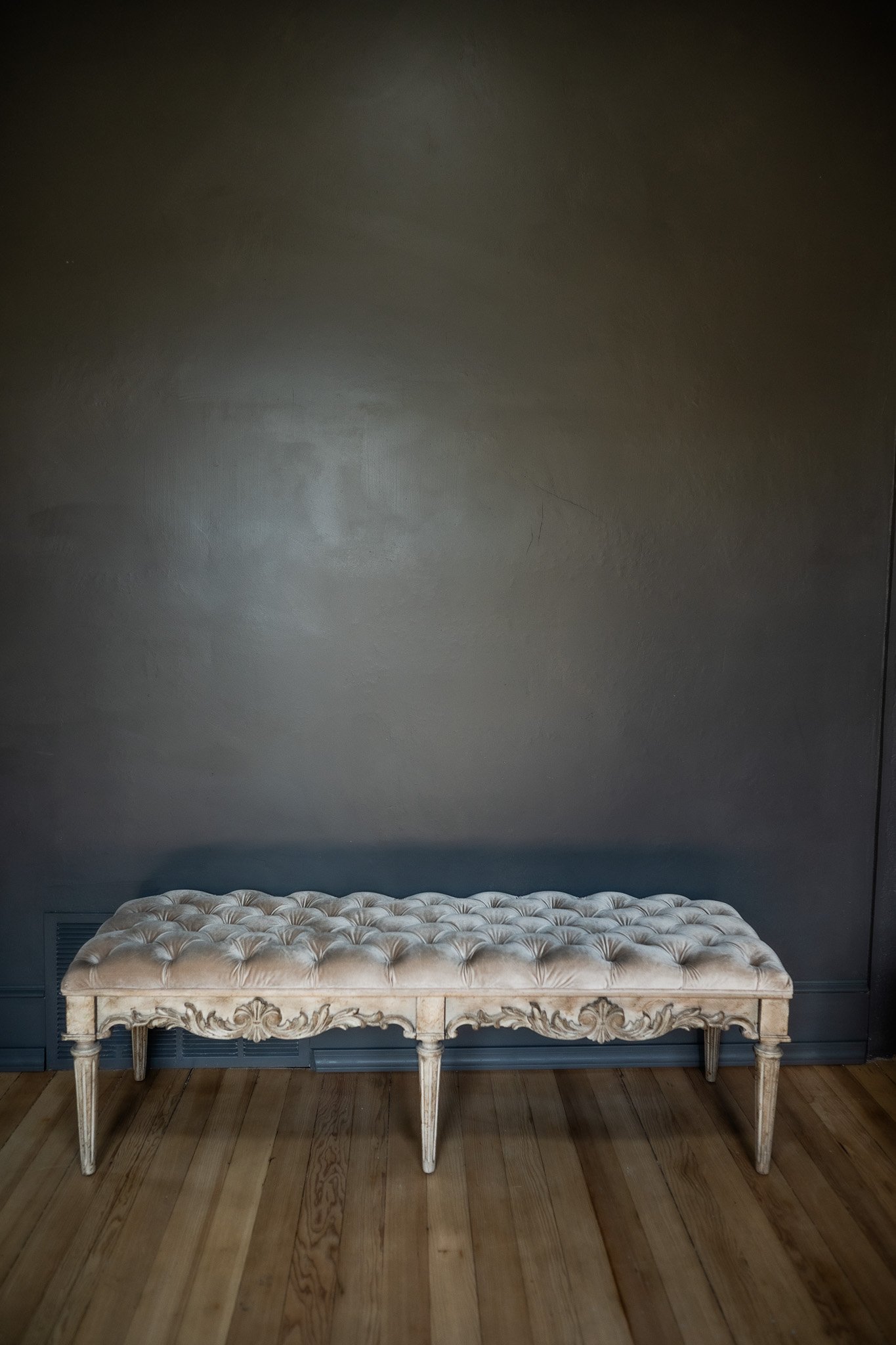 An elegant vintage-style beige tufted bench with carved wooden legs placed against a dark gray wall on a wooden floor.