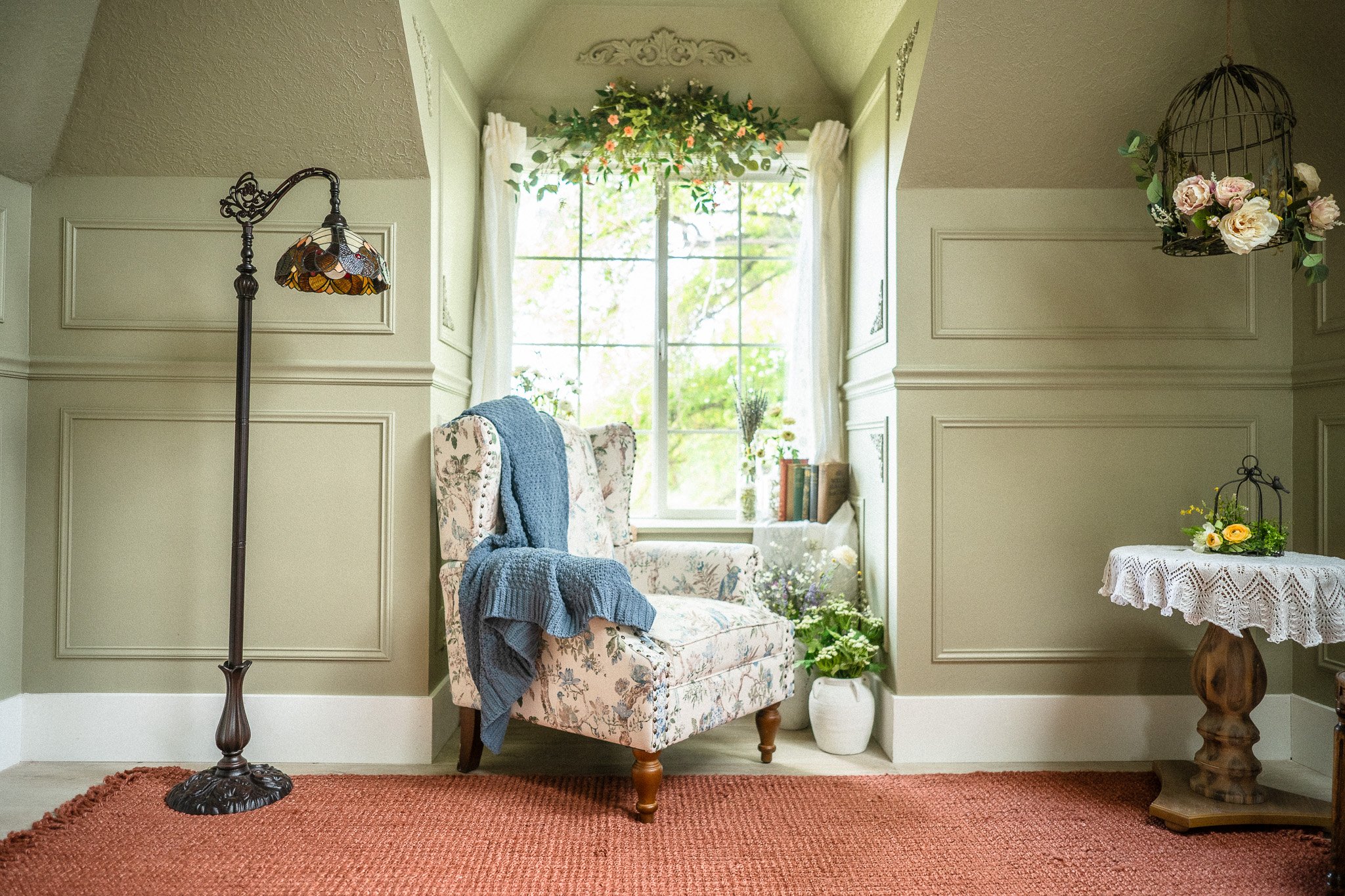 Cozy living room corner with a floral upholstered armchair draped with a blue knitted blanket, a window with white curtains and floral greenery above, a white pot with plants on the window ledge, a small side table with a lace cloth and flower arrang