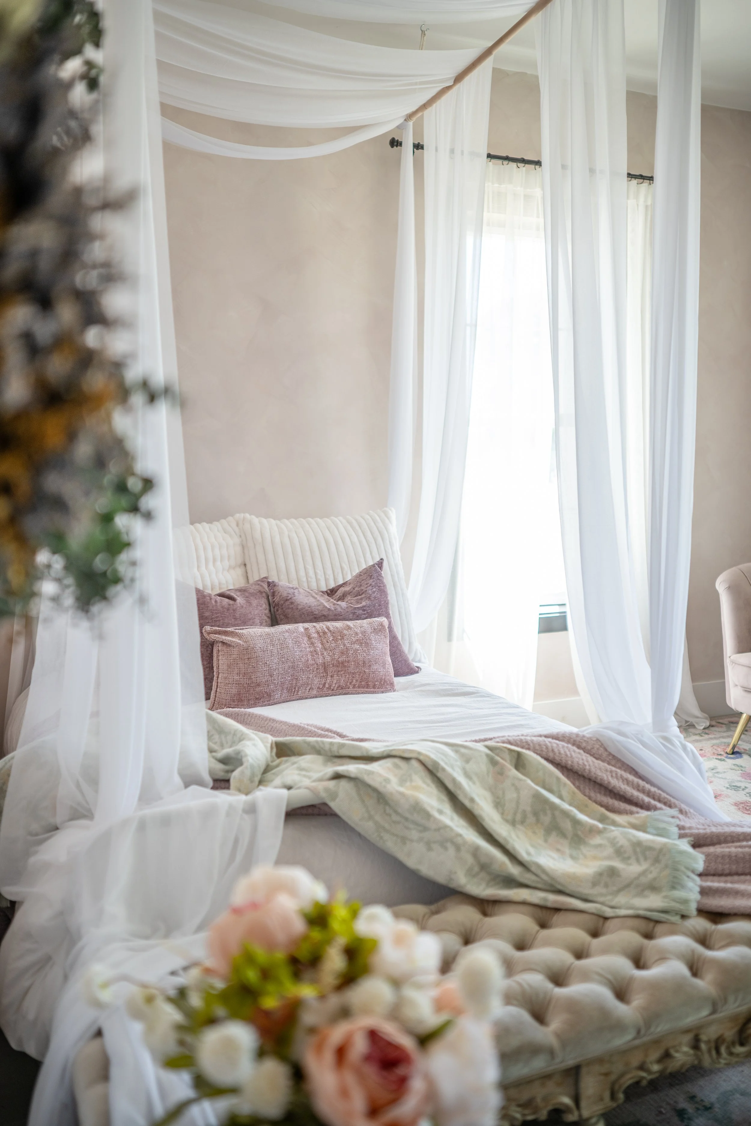Elegant bedroom with a four-poster bed draped in sheer white curtains, soft pink and beige pillows, and a floral blanket. In the foreground, a blurred bouquet of pale pink and white flowers is visible.