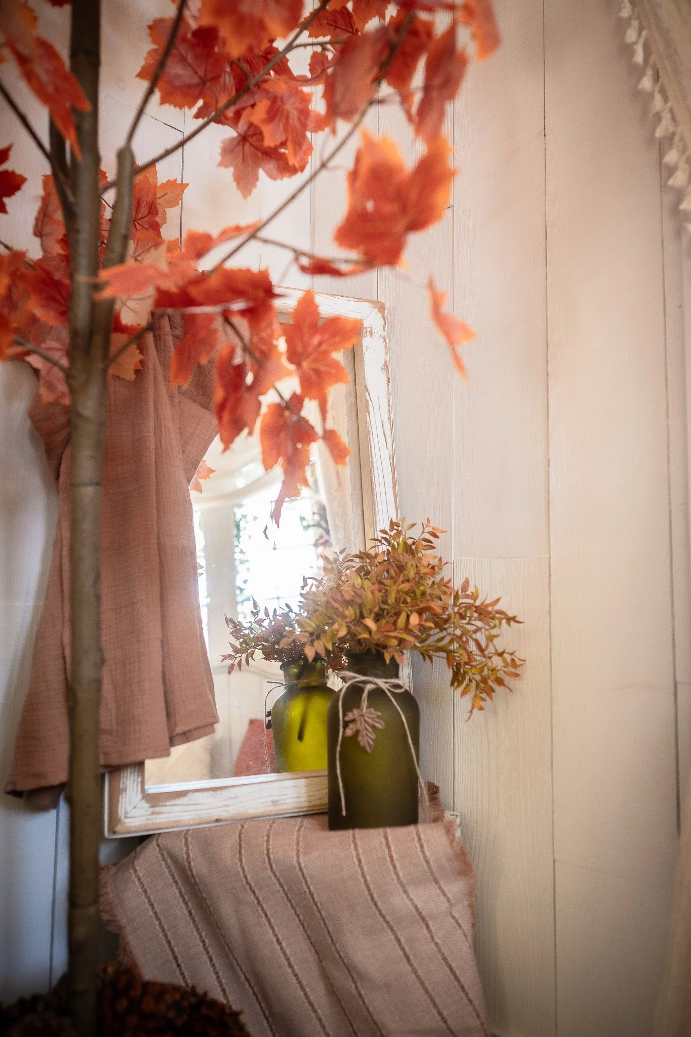 Decorative interior with orange leaves on a tree branch, a mirror reflecting a window, a beige striped cloth, and green vases with brown and orange foliage.