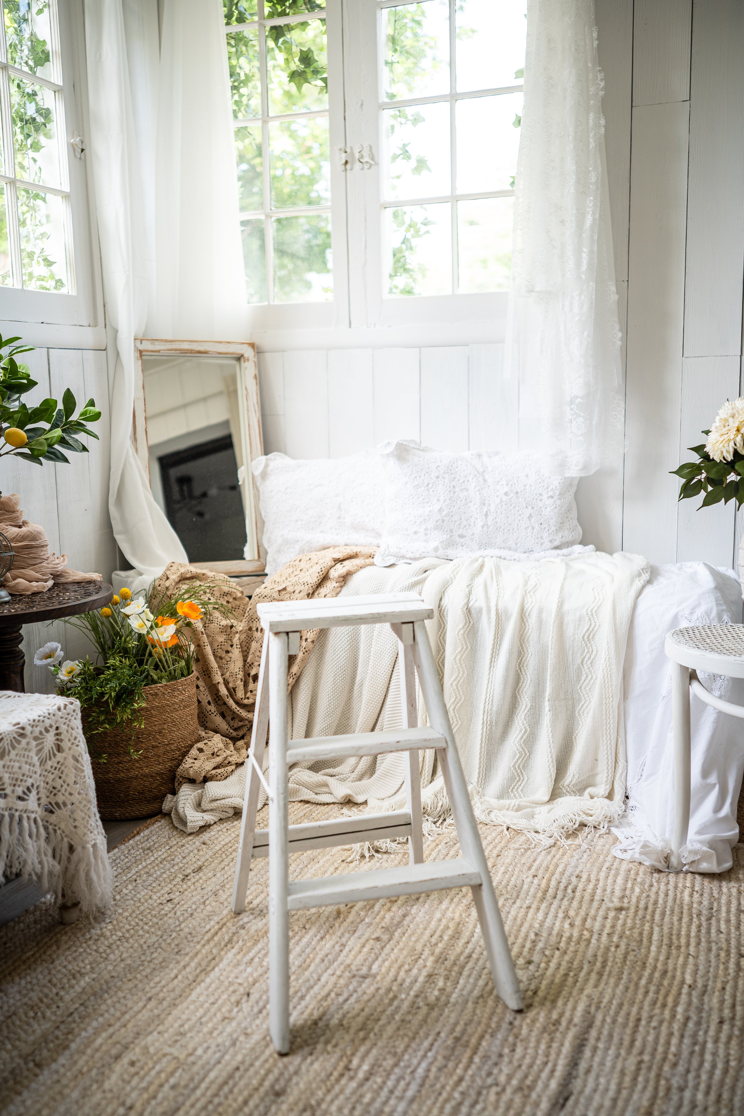 A cozy, bright room with white wooden panel walls, windows with white lace curtains, and a window seat with white pillows and textured blankets. There is a large mirror leaning against the wall, a potted plant with yellow flowers, and a white painted
