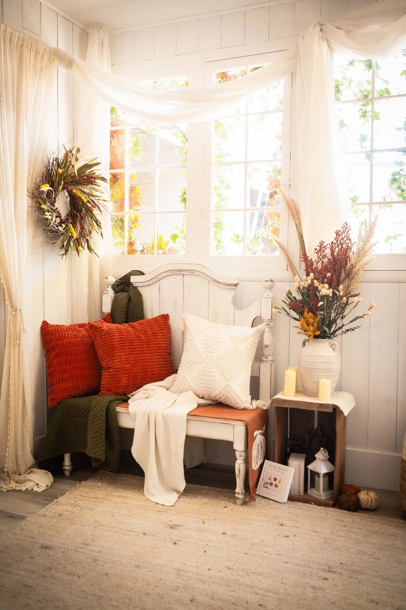 Cozy corner with white wooden bench, orange and white pillows, draped white blankets, a vase with dried flowers, candles, and autumnal decor, near a window with cream curtains.