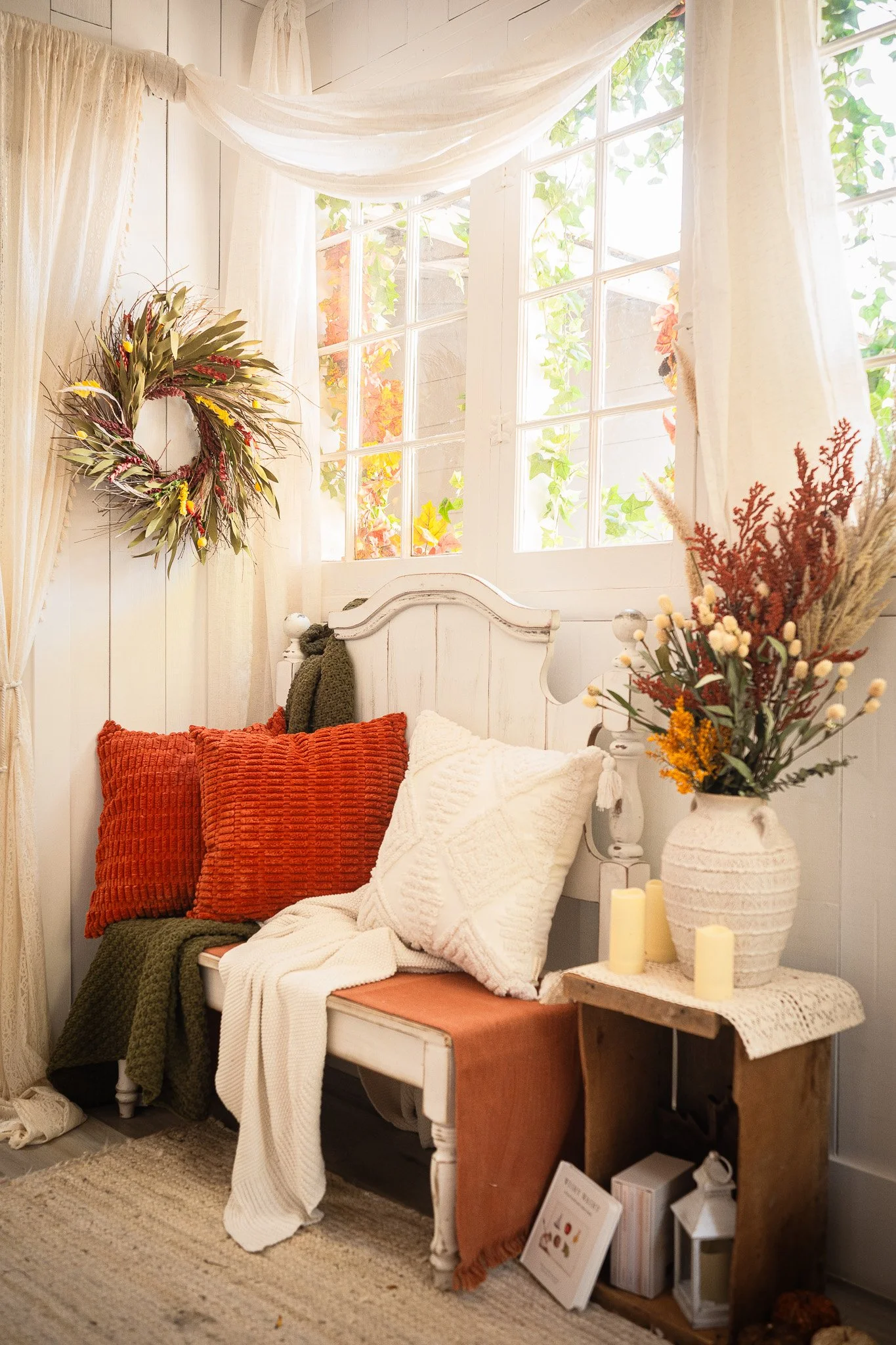 Cozy corner with white wooden bench, orange pillows, and a white blanket, next to a small rustic table with a large beige vase of dried flowers and yellow candles, beside a window with flowy curtains and autumn foliage.