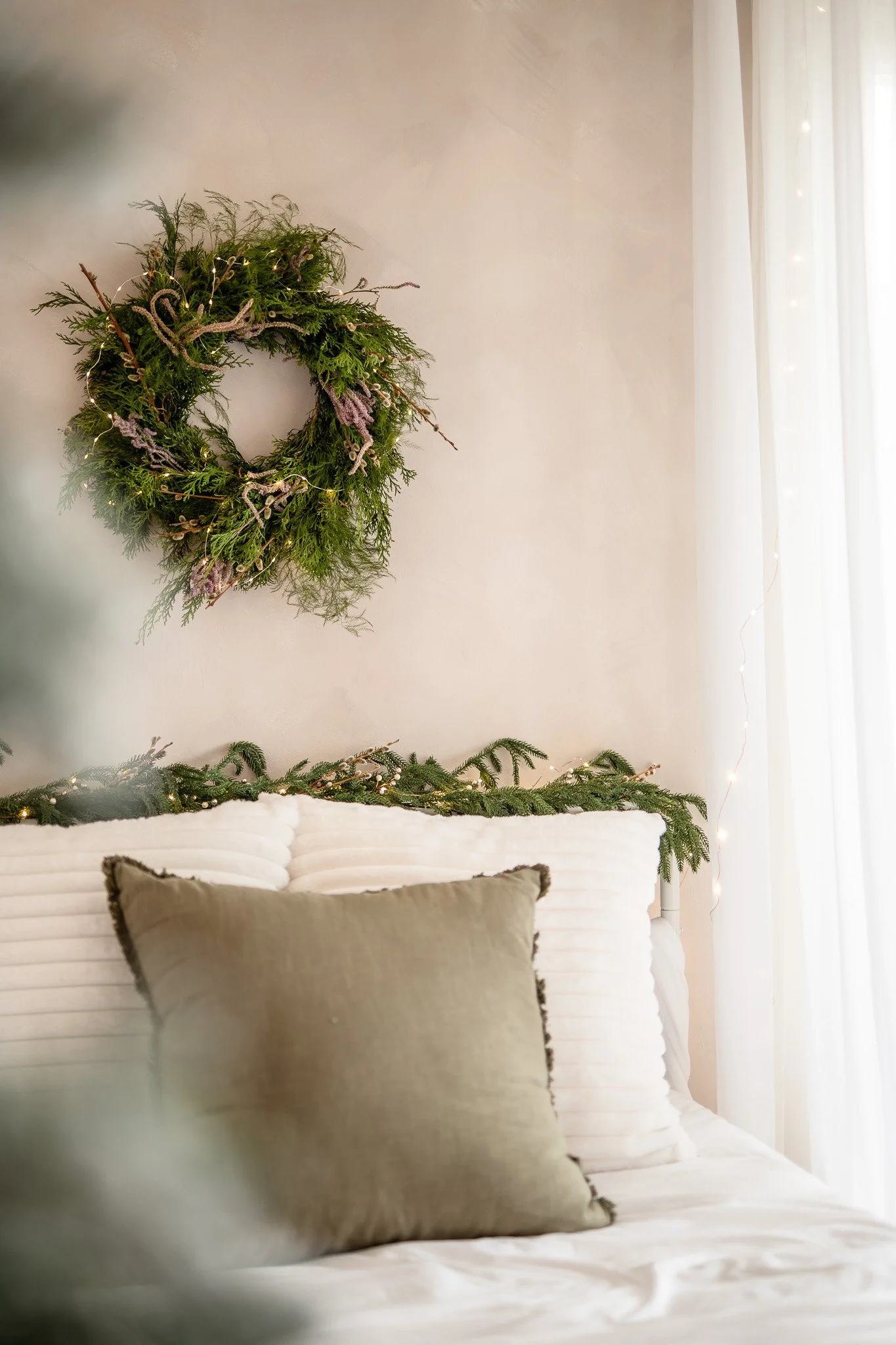 A Christmas wreath hanging on a beige wall behind a bed decorated with white blankets, beige pillows, and a green garland along the headboard, illuminated by string lights and in front of sheer white curtains.