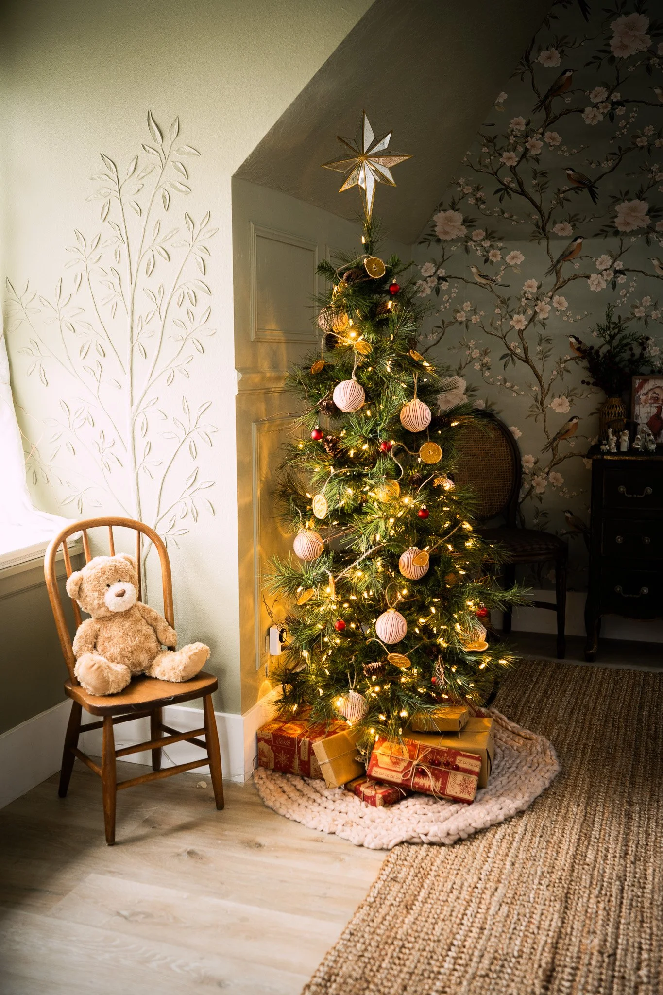A decorated Christmas tree with ornaments and lights, topped with a star, surrounded by wrapped gifts. To the left, a teddy bear sits on a wooden chair near a window. The room features a floral wallpapered wall.
