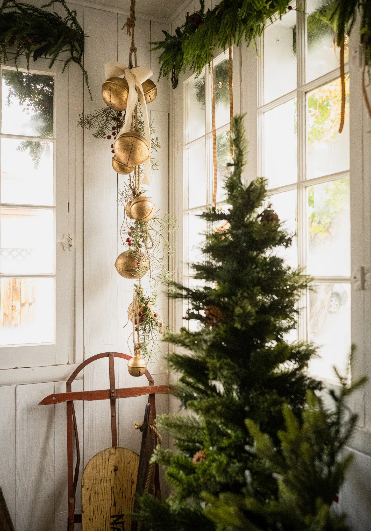 A corner of a room decorated for Christmas with a small Christmas tree, a vintage wooden sleigh, and a hanging garland of gold ornaments and greenery, all illuminated by natural light from large window panes.