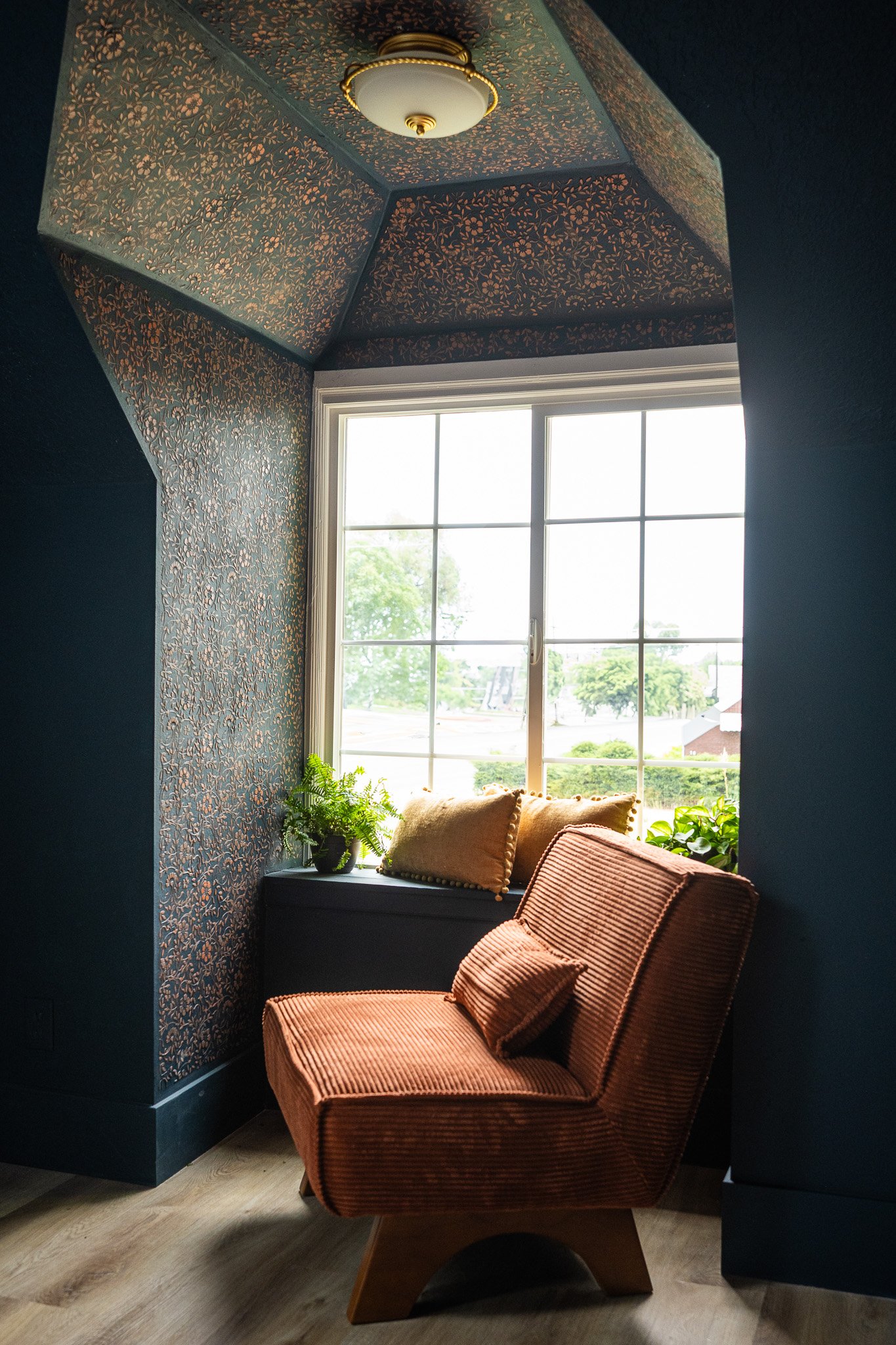 A cozy reading nook with an orange velvet armchair with a matching pillow, a window seat with two cushions, and potted plants on the window ledge, surrounded by wallpapered and painted walls.