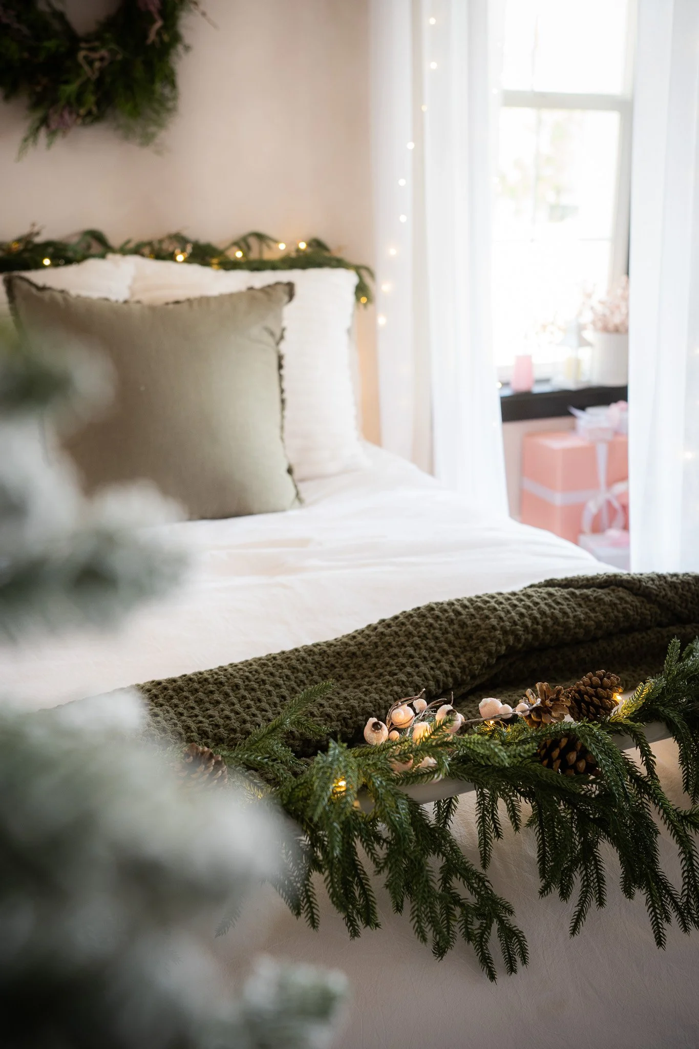 A cozy bedroom decorated for Christmas with a bed featuring white and gray pillows, a dark green textured blanket, and garland with pine cones and lights. In the background, a window with sheer white curtains reveals pink gift boxes and candles on th