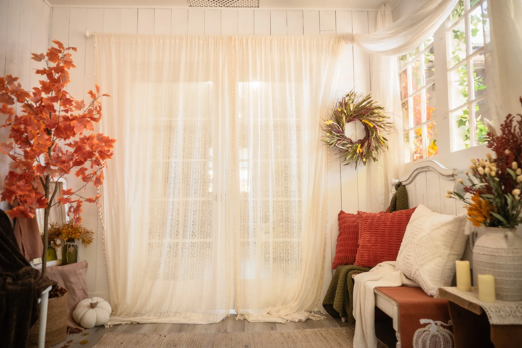 Cozy room with beige curtains, orange and white pillows on a white bench, and fall decorations including a wreath and potted plants with autumn leaves.