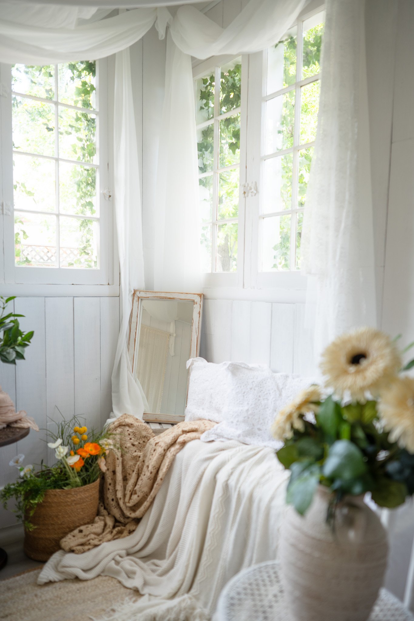 A cozy corner of a room with white wooden walls and large windows with white curtains. There are various plants and flowers, a leaning mirror, and a beige blanket on a cushioned white bench.