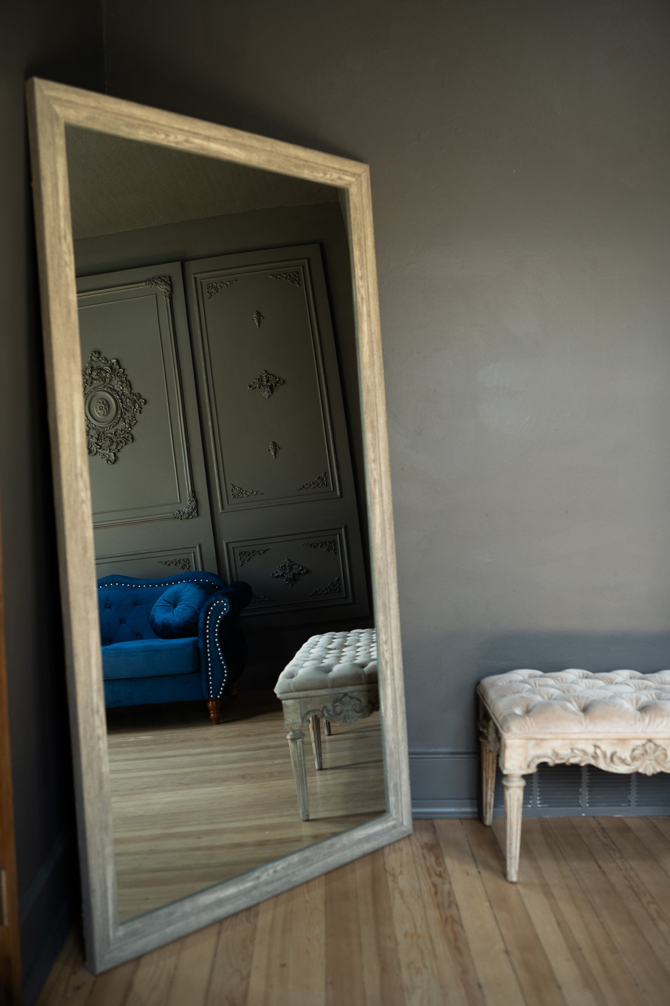 Reflection of a blue tufted velvet sofa, a beige quilted bench, and ornate wall decor visible in a large standing mirror in a room with wooden floors and gray walls.
