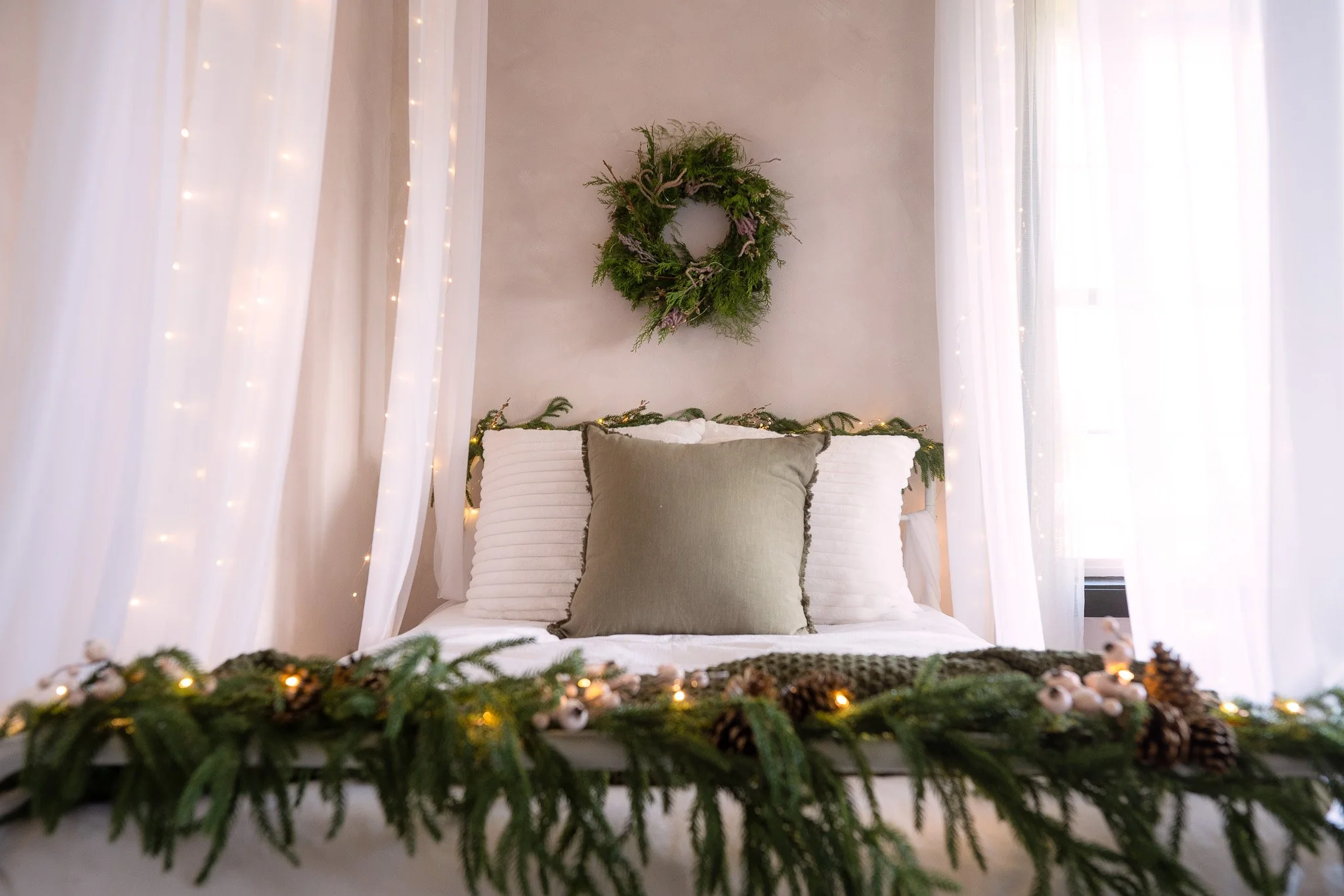 A cozy bedroom decorated with holiday greenery, pinecones, and string lights. The bed has white and beige pillows and a green throw pillow, with a wreath hanging on the wall above. Sheer white curtains frame the window and add softness to the scene.