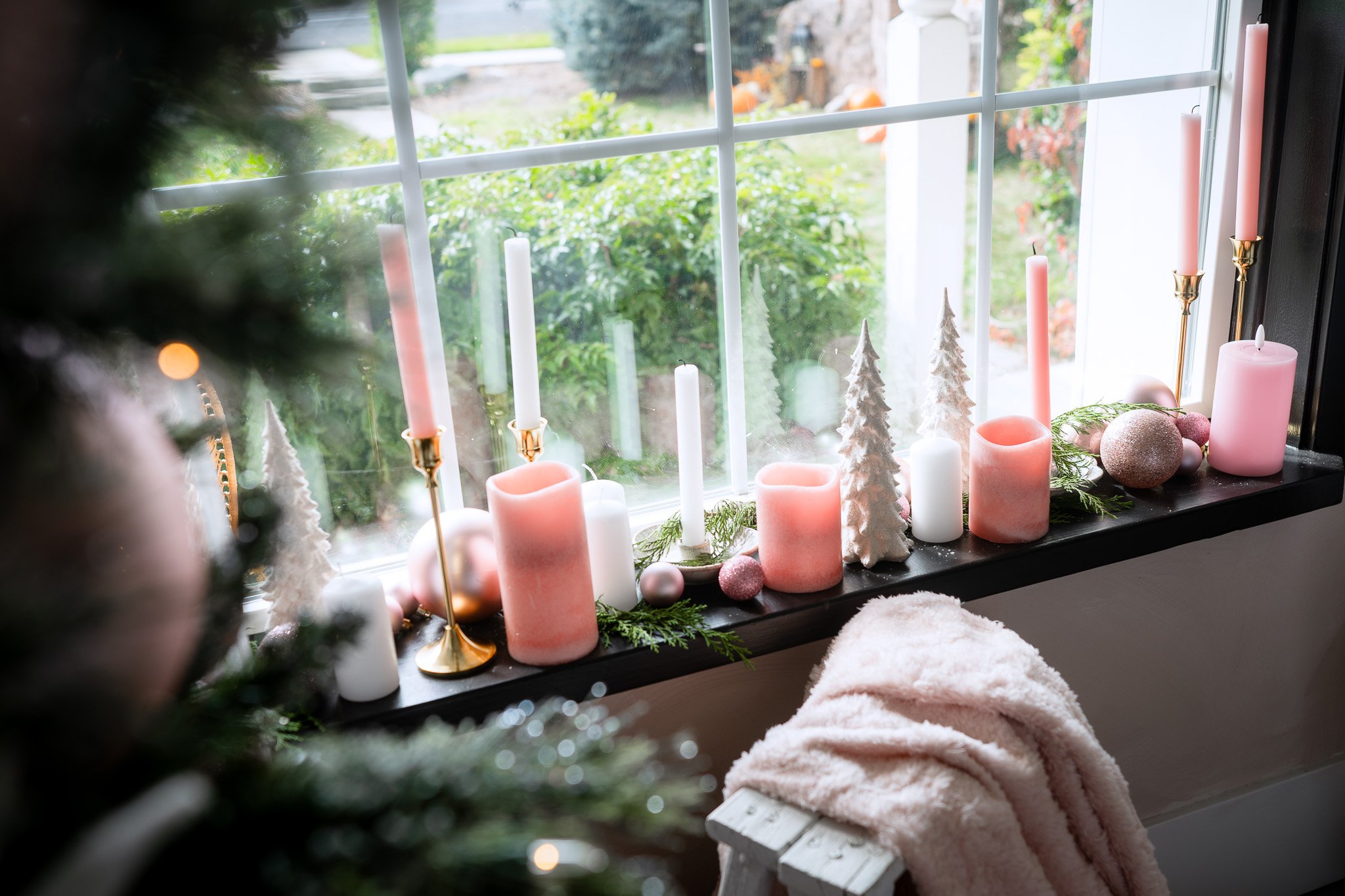 Decorative Christmas window display with pink, white, and gold candles, pastel ornaments, small Christmas trees, and greenery, viewed from inside the house.