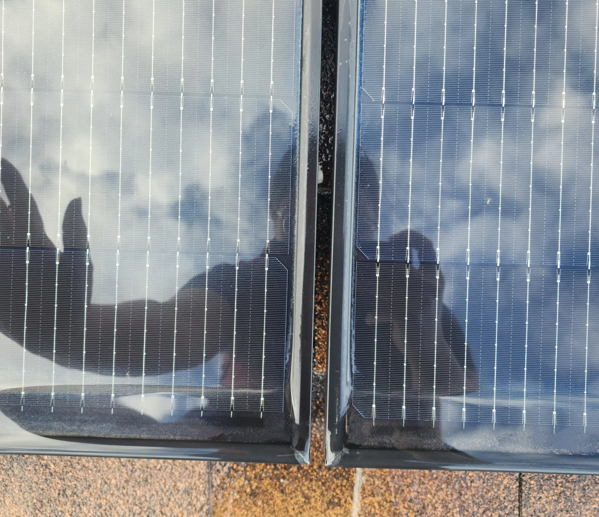Close-up of two solar panels with reflections of a person's silhouette and clouds, mounted on a rooftop.