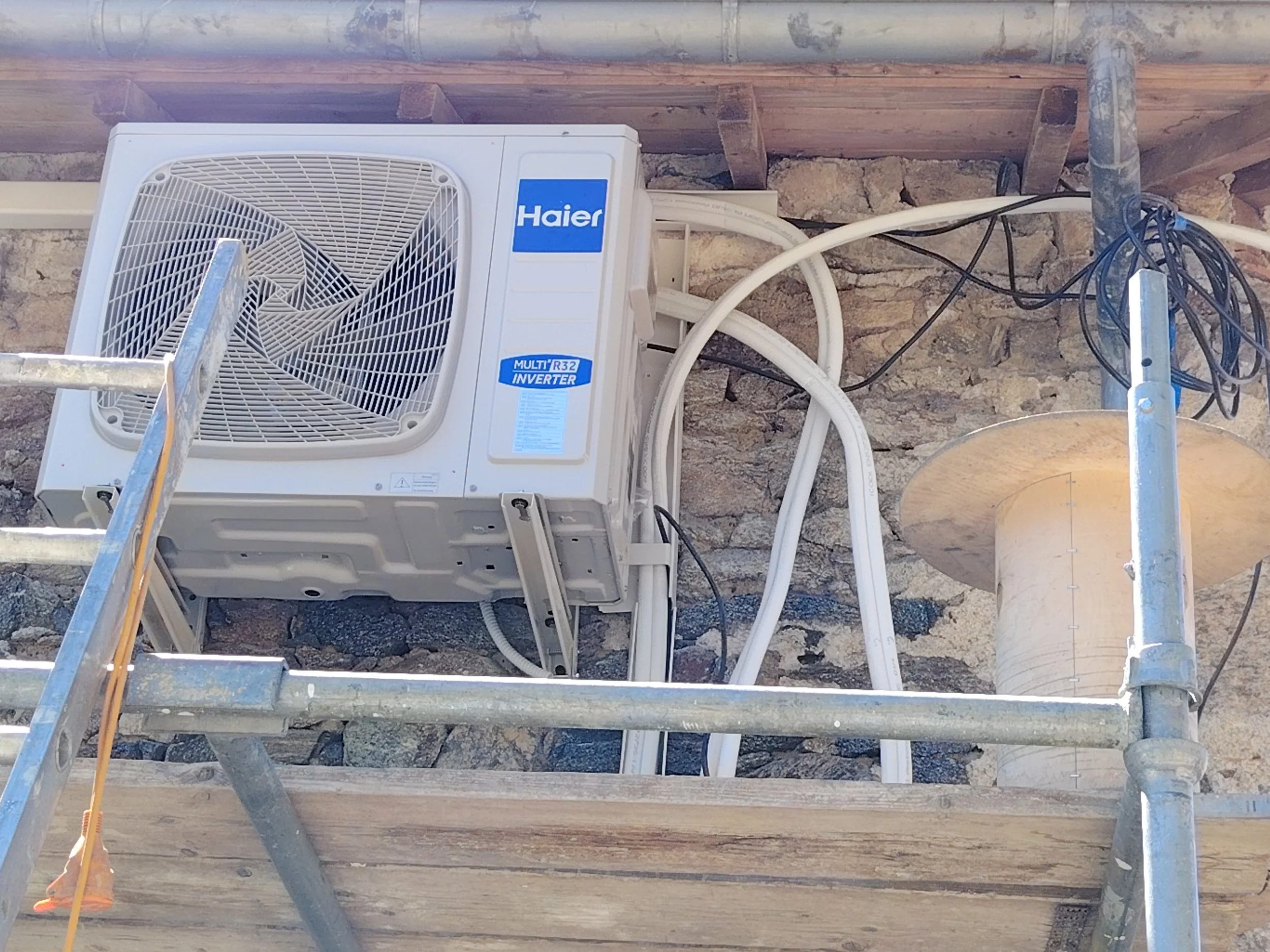Air conditioning unit on a construction site with scaffolding and electrical wires against a stone wall.