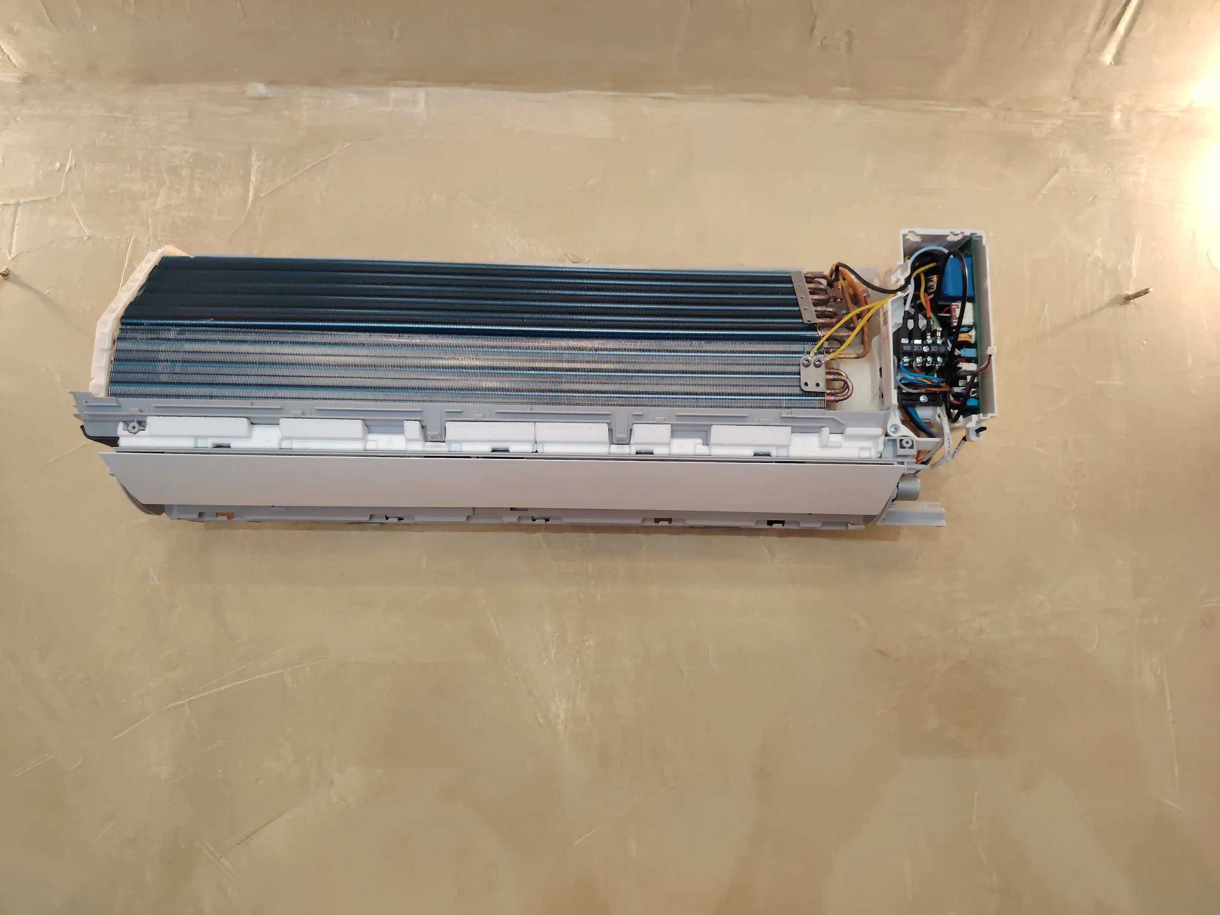 Disassembled air conditioning indoor unit with visible fan, electronic components, wiring, and heat exchange coil, lying on a beige floor.