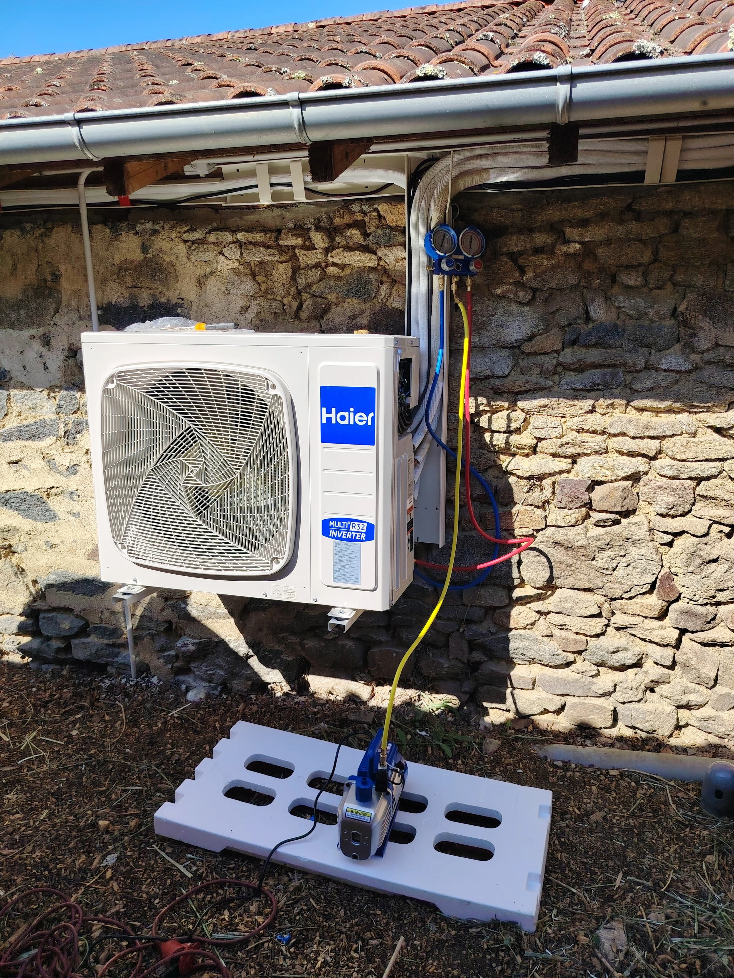 Outdoor view of a Haier air conditioning unit mounted on a stone wall with refrigerant lines connected to gauges on a worktable.