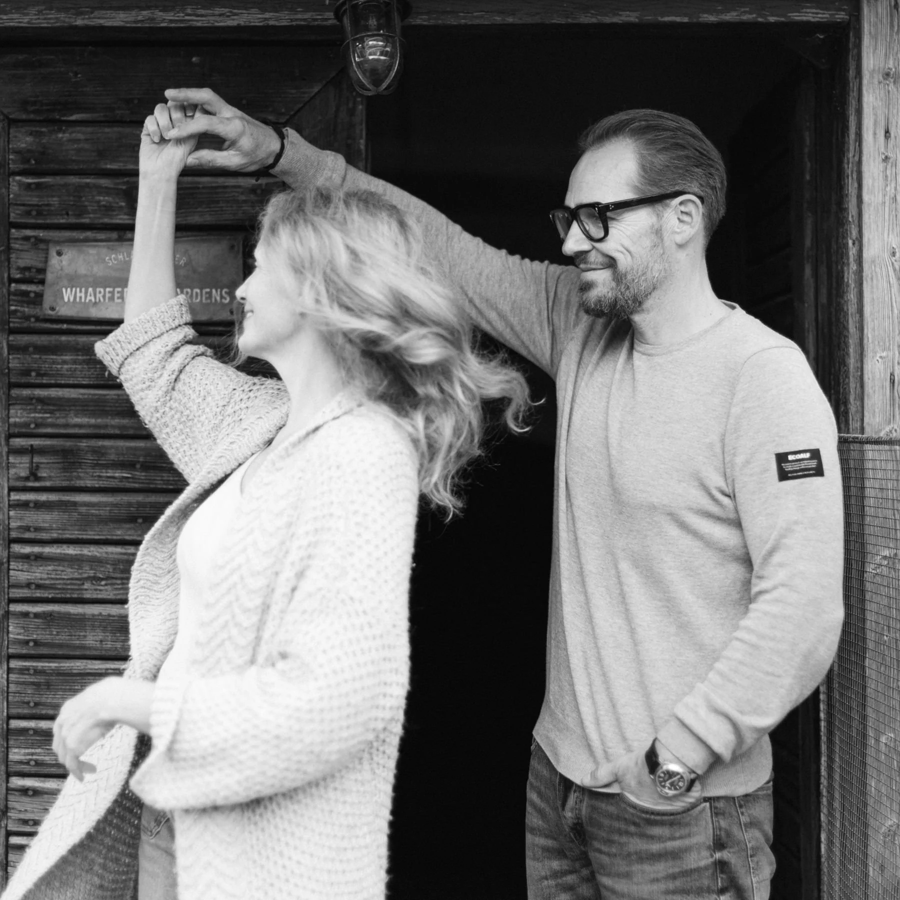 Ein Mann mit Brille und eine Frau mit Locken posieren in einem schwarzen und weißen Foto; er berührt ihre Arme verspielt, vor einer Holzwand im Freien.