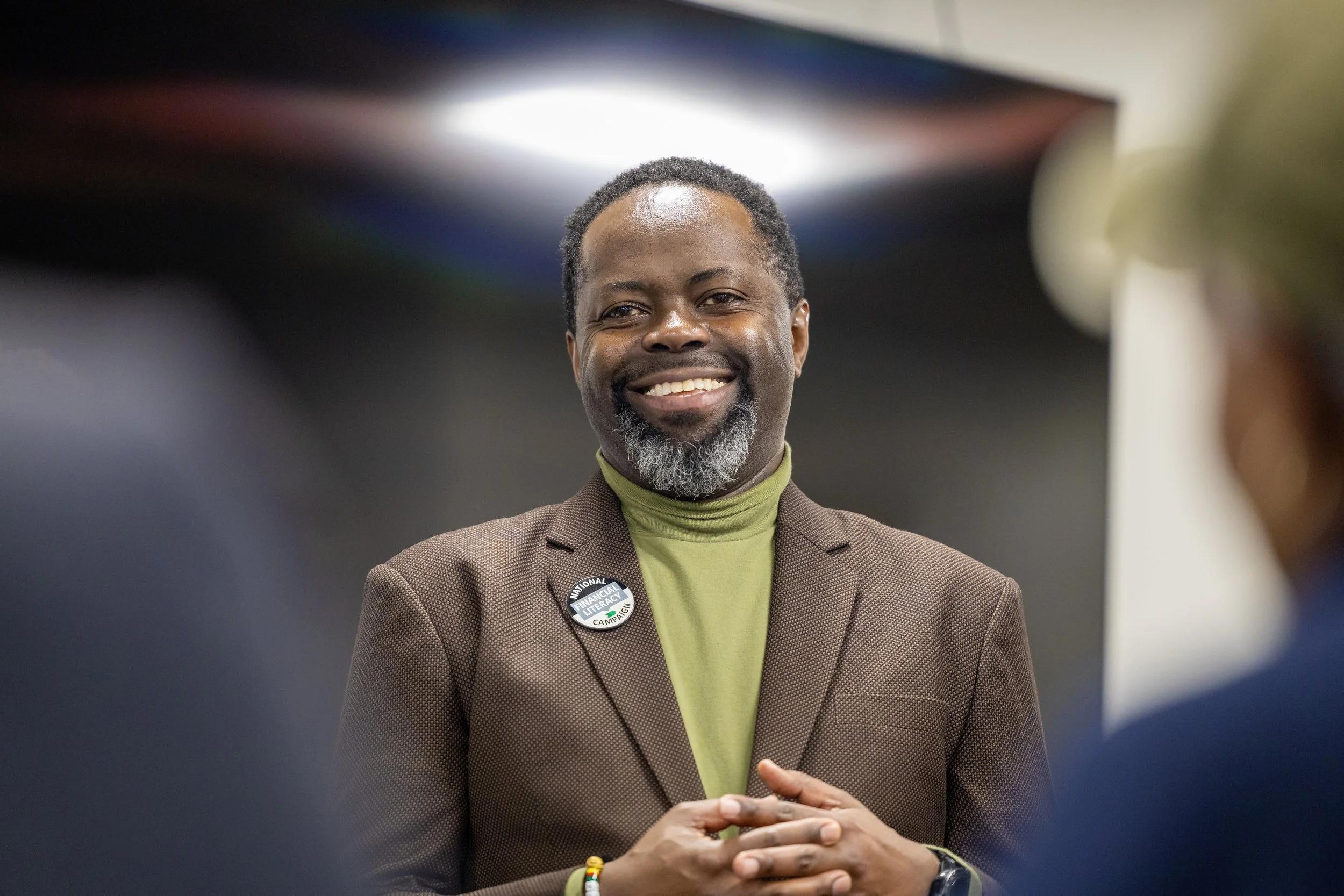 A man with a beard and mustache, wearing a brown blazer and a green turtleneck, smiling and holding his hands together, standing in front of a blurred background.