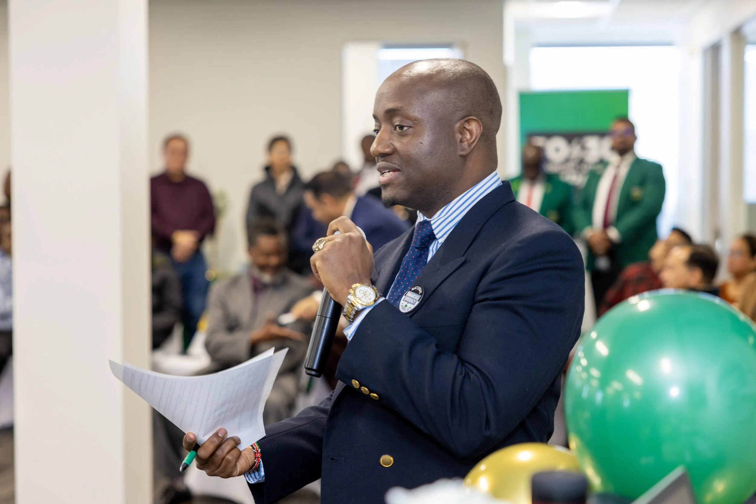 A man in a navy suit speaking into a microphone during a conference, with attendees seated and standing in the background.