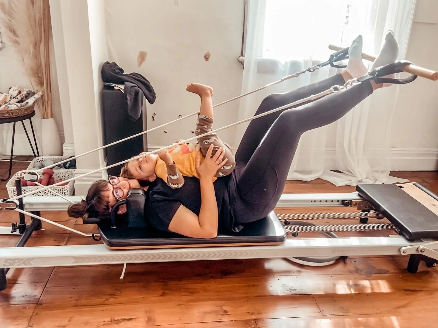 A woman and her child are lying on a Pilates reformer machine, both smiling, in a The Reformer Pilates room with hardwood floors and white curtains.