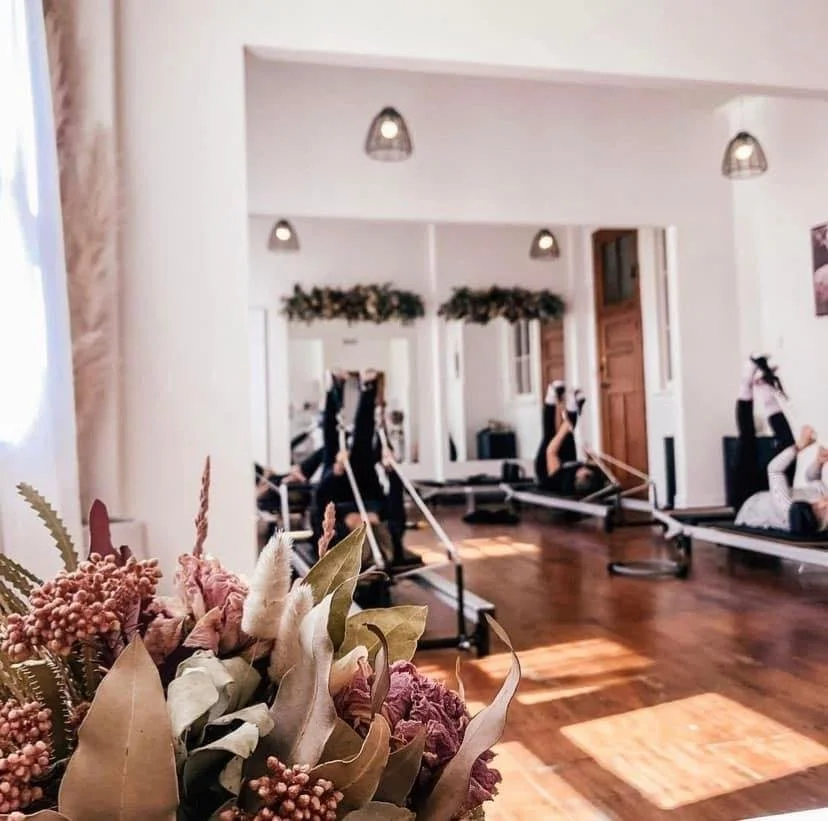 A group of people in a Reformer Pilates studio exercising on reformer machines, with a bouquet in the foreground.
