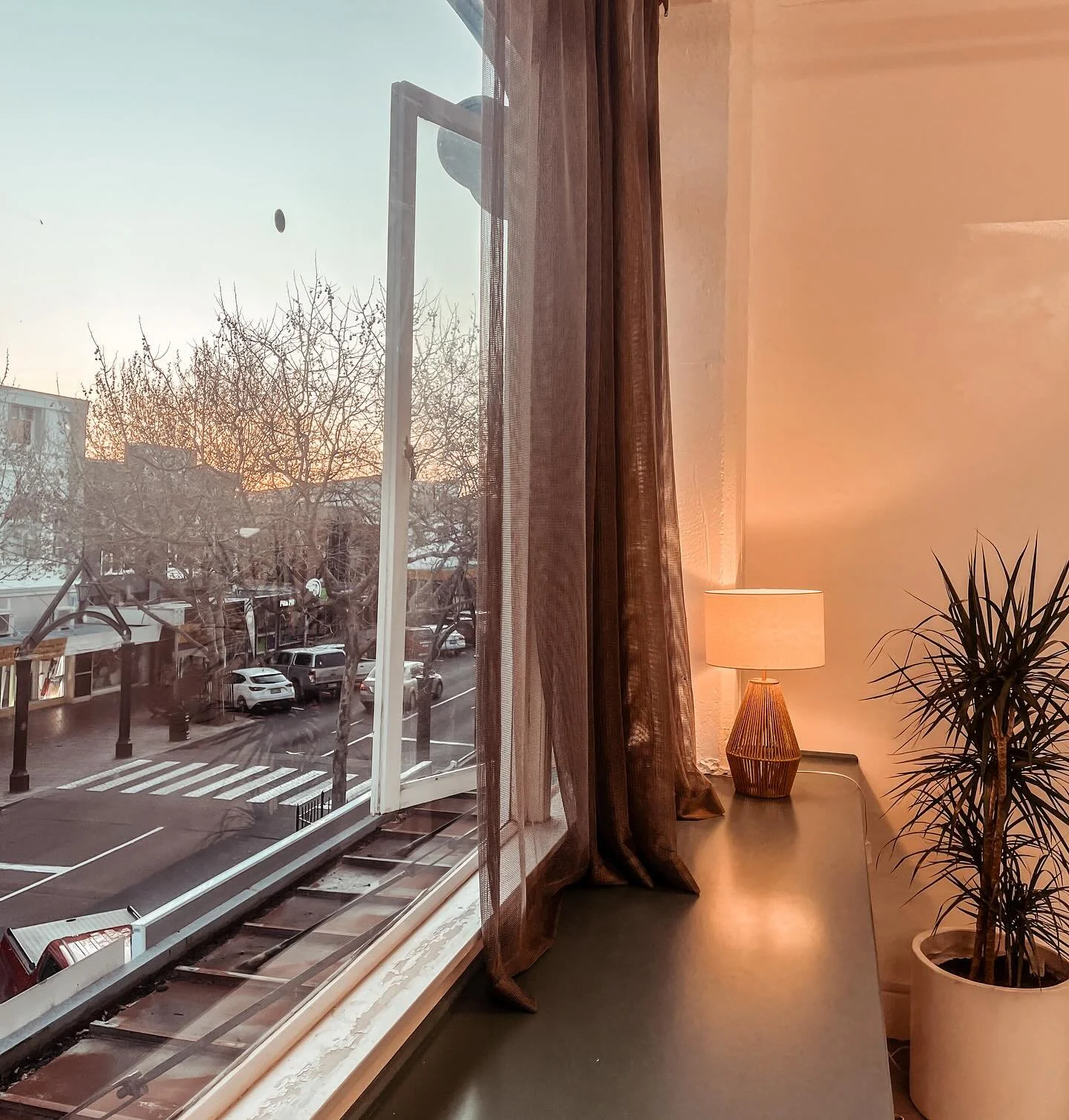 Interior scene showing the window at Reformer Pilates with open blinds and curtains, a lamp, and a potted plant on a table, with the street visible outside during sunset.