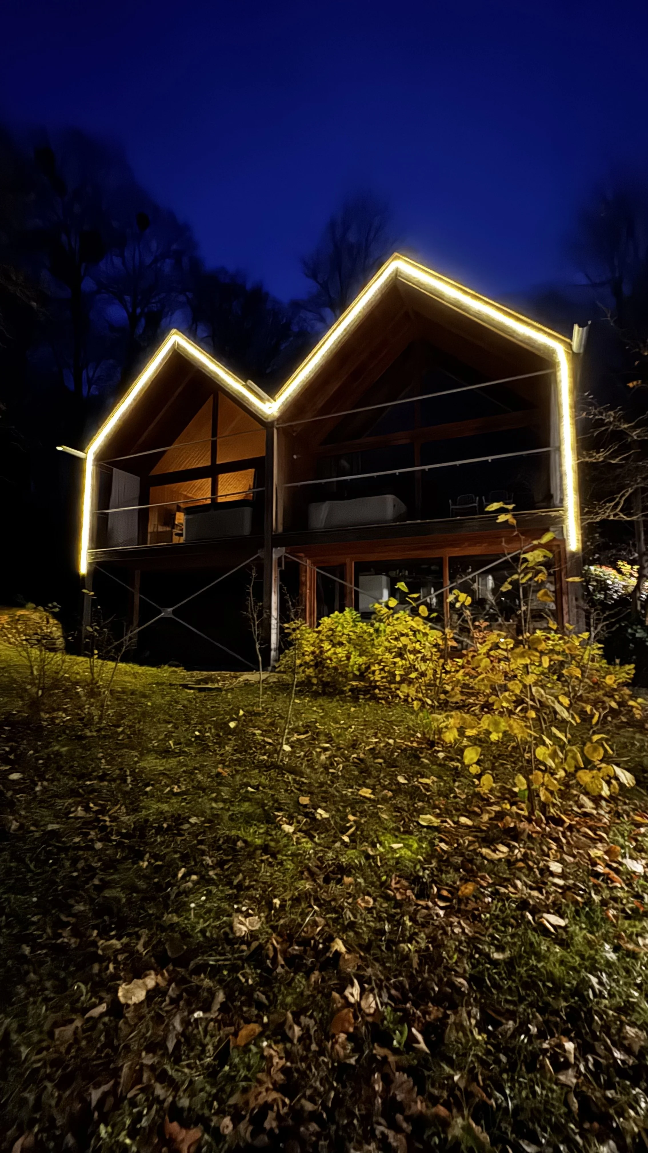 A modern house with illuminated outline at night, situated on a grassy terrain with fallen leaves.