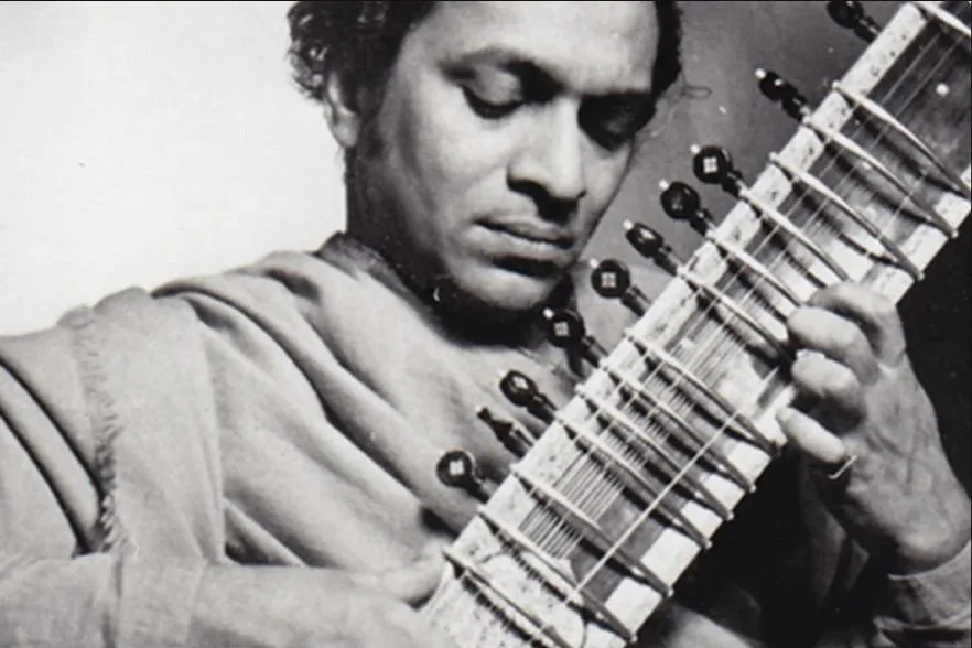 Black and white photo of a young man with dark curly hair, playing a sitar guitar.