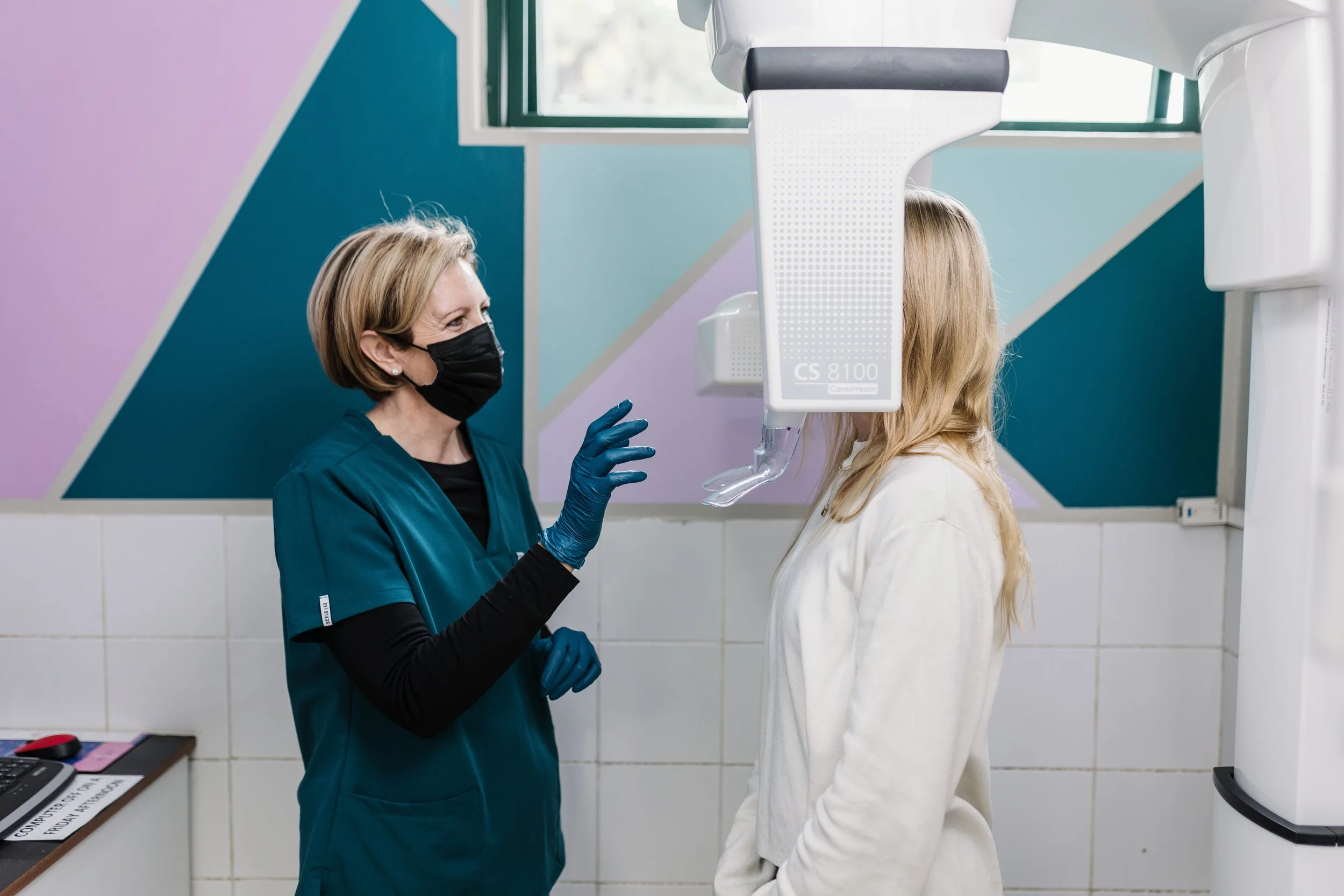 A healthcare professional wearing a black face mask and teal scrubs is speaking with a patient inside a medical or dental office, with a X-ray machine overhead.