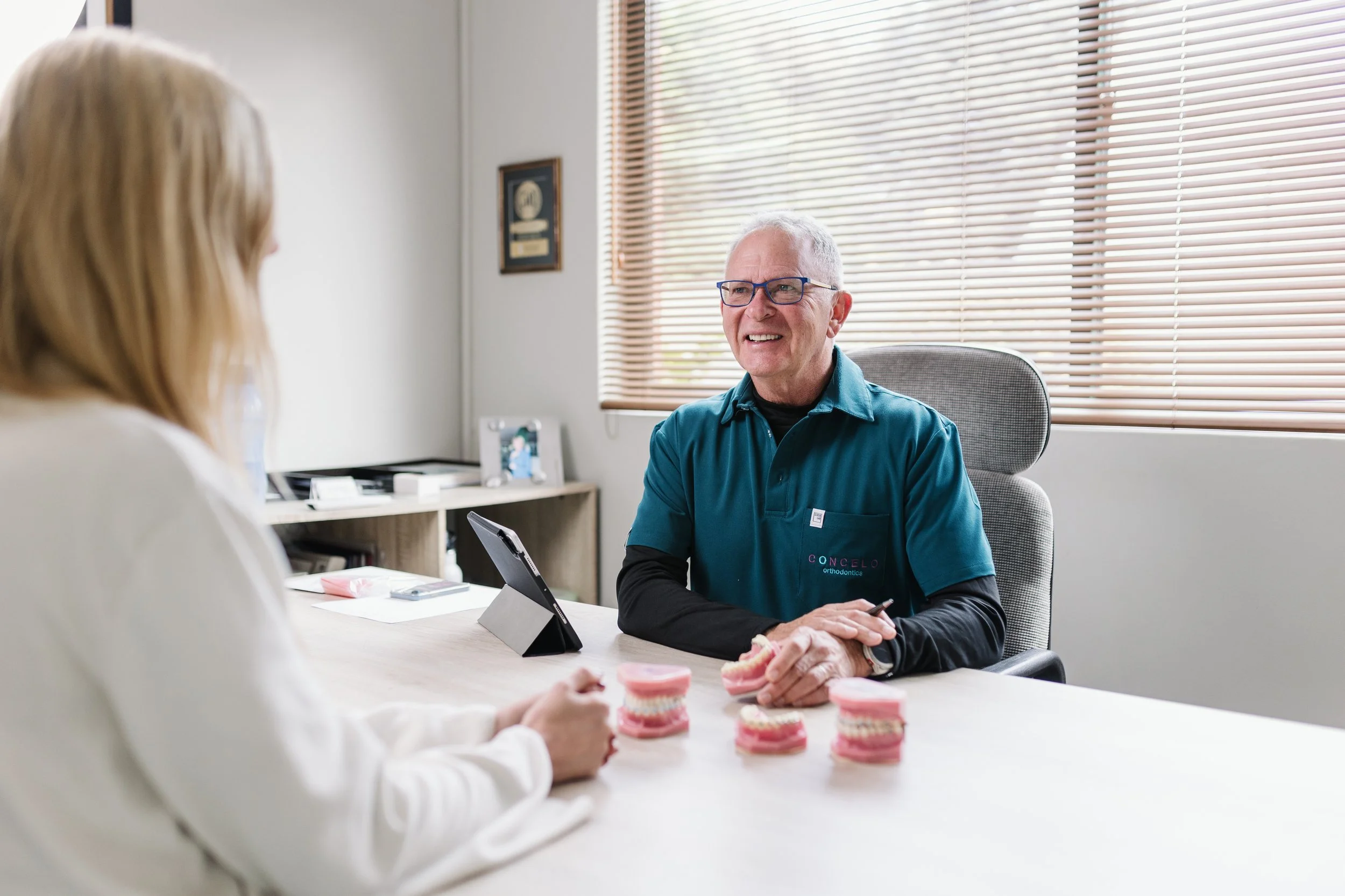 A male orthodontist wearing glasses and a teal shirt with an 'Concelos Orthodontics' logo, talking to a patient in a dental office. The patient, with blonde hair, is partly visible and facing away from the camera. On the table, there are dental models and a tablet. The office has large windows with blinds and a framed certificate on the wall.