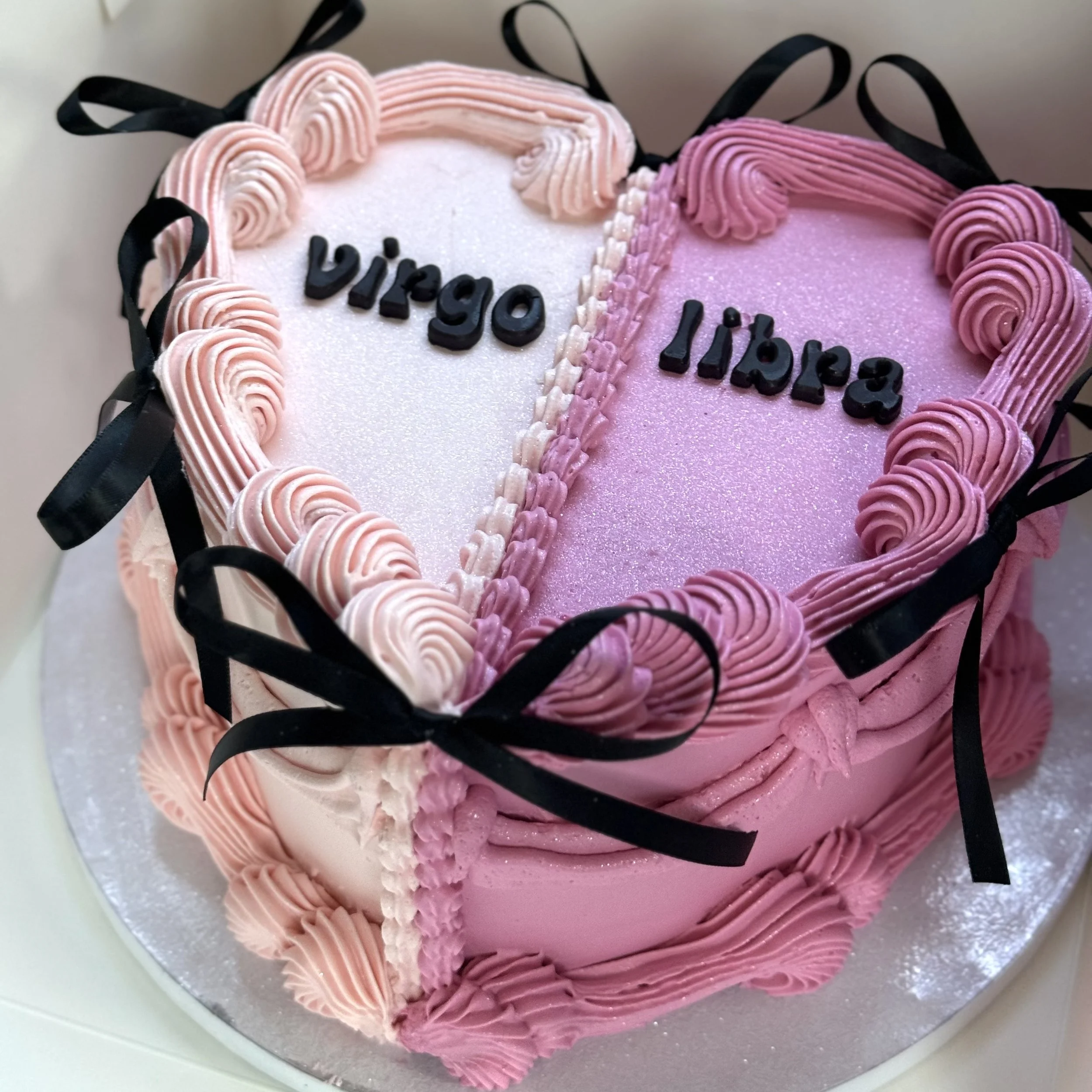 Pink heart-shaped cake decorated with pink and white frosting, black ribbons, and the names 'Virgo' and 'Libra' written on it.
