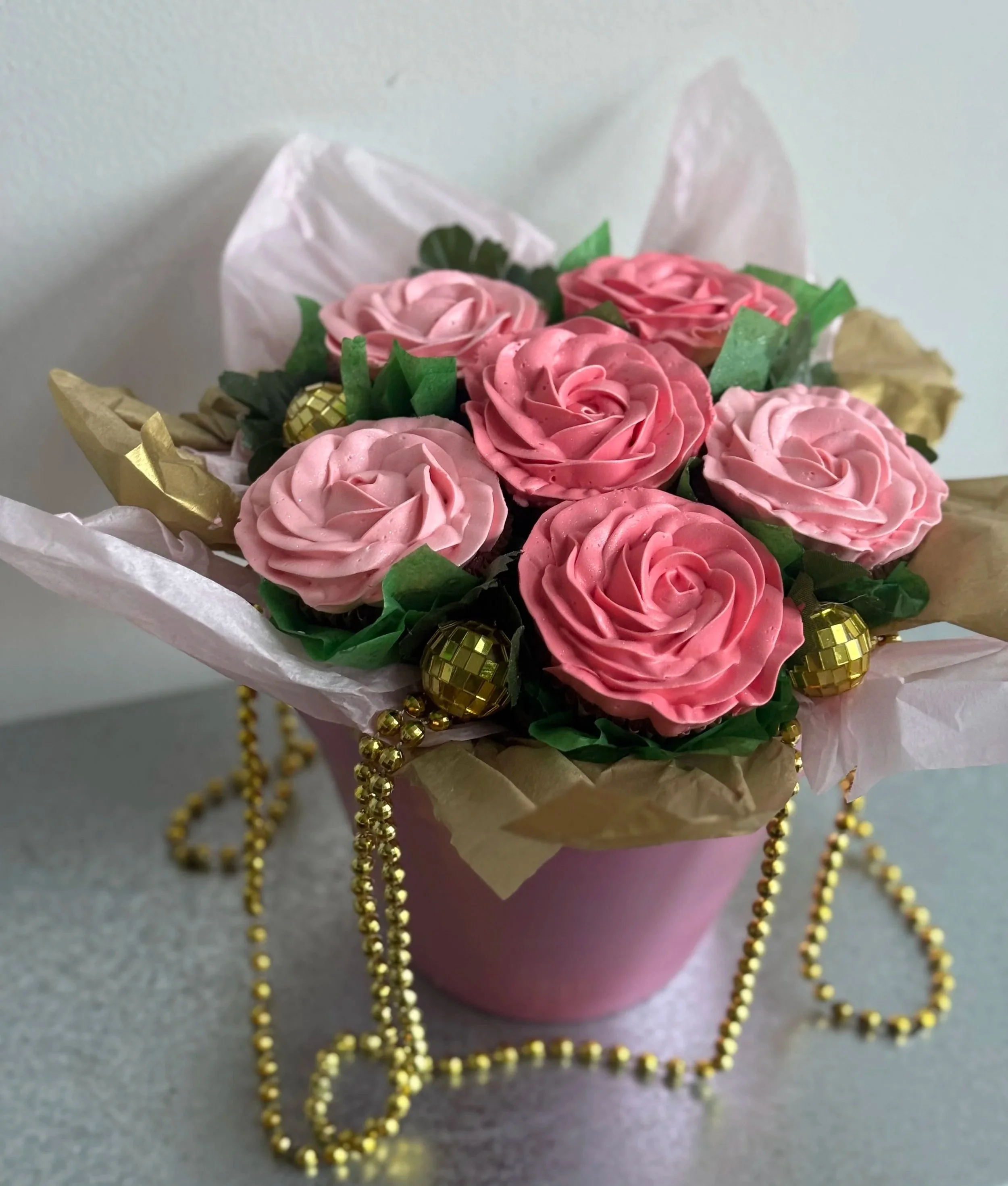 A pink flower arrangement with artificial roses, green leaves, gold accents, and a gold bead necklace in a pink pot on a gray surface.