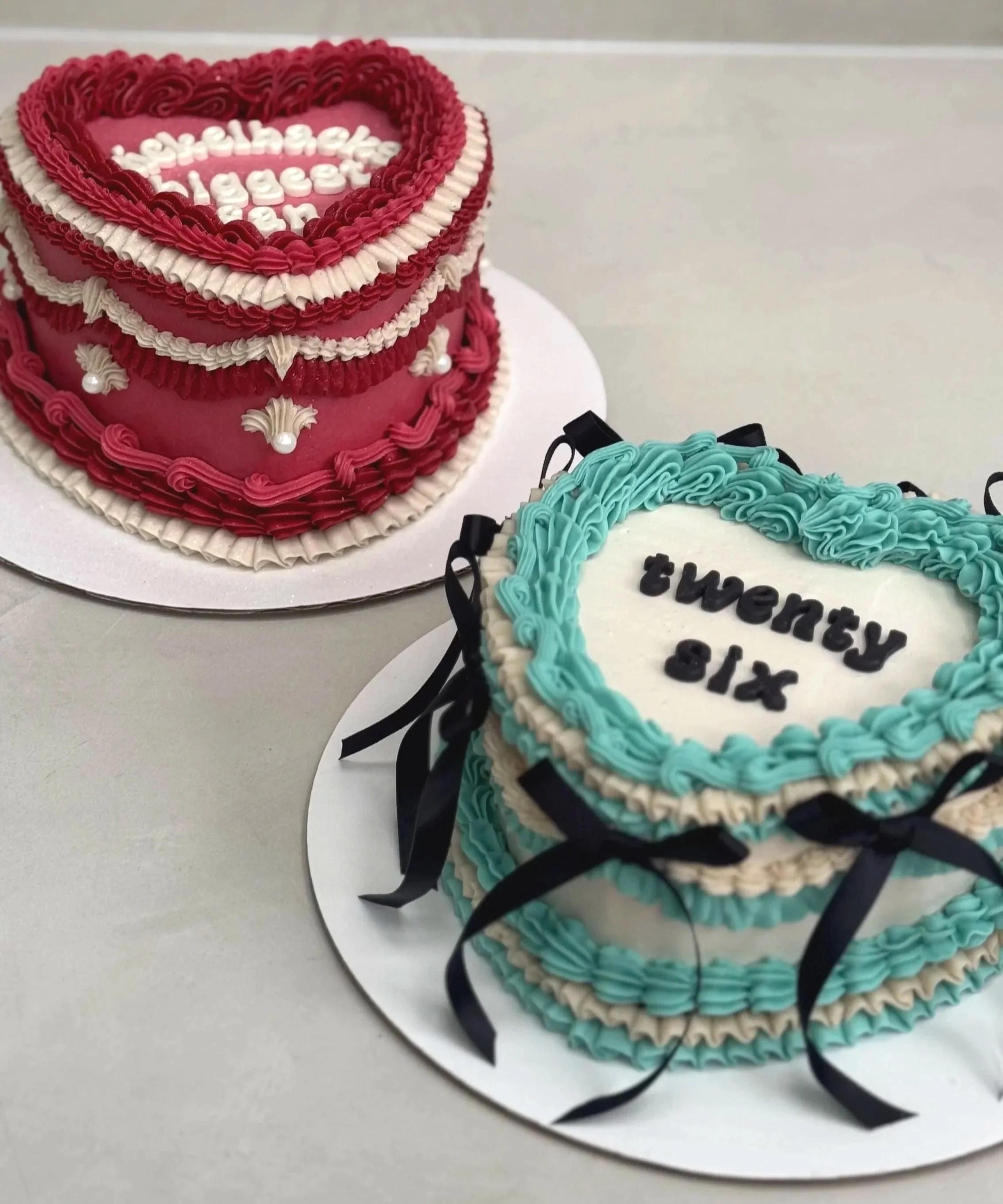 Two decorated cakes for a celebration. One cake is heart-shaped with pink and white frosting, featuring the words "Happy Anniversary" and decorative piping. The other cake is rectangular with blue and white frosting, with the words "Twenty Six" writt