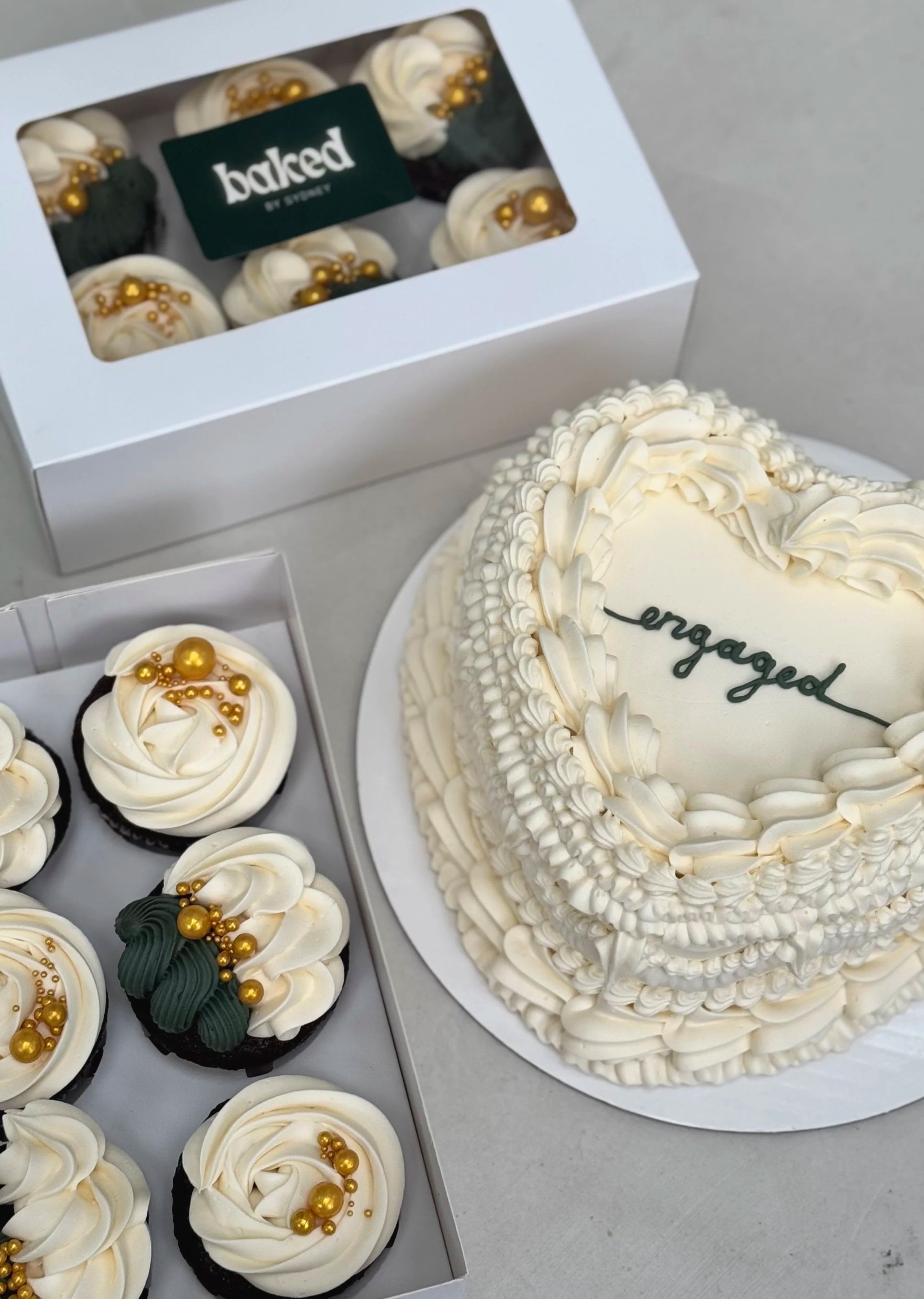 Celebration cake with white frosting and the word 'engaged' written on it, decorated with piped frosting, and a box of cupcakes with white frosting, gold edible beads, and dark green frosting decorations.
