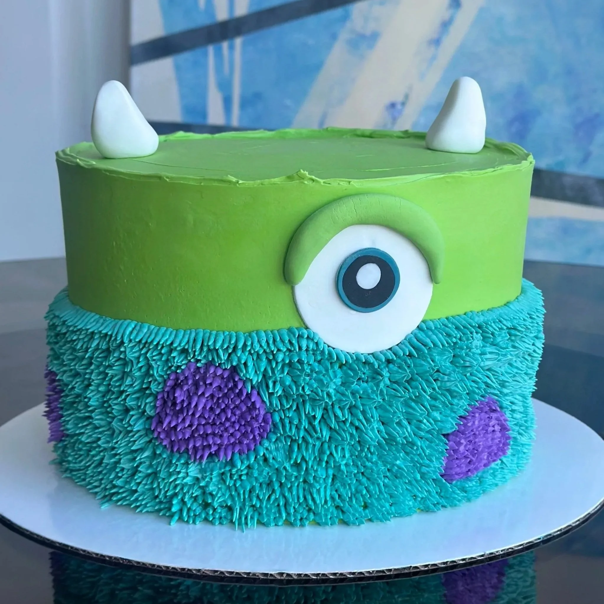 A two-layer birthday cake decorated with a monster theme. The top layer is green with two white horn-like decorations. The side of the bottom layer is covered in blue frosting with purple spots, and features a large green eye with white and black det