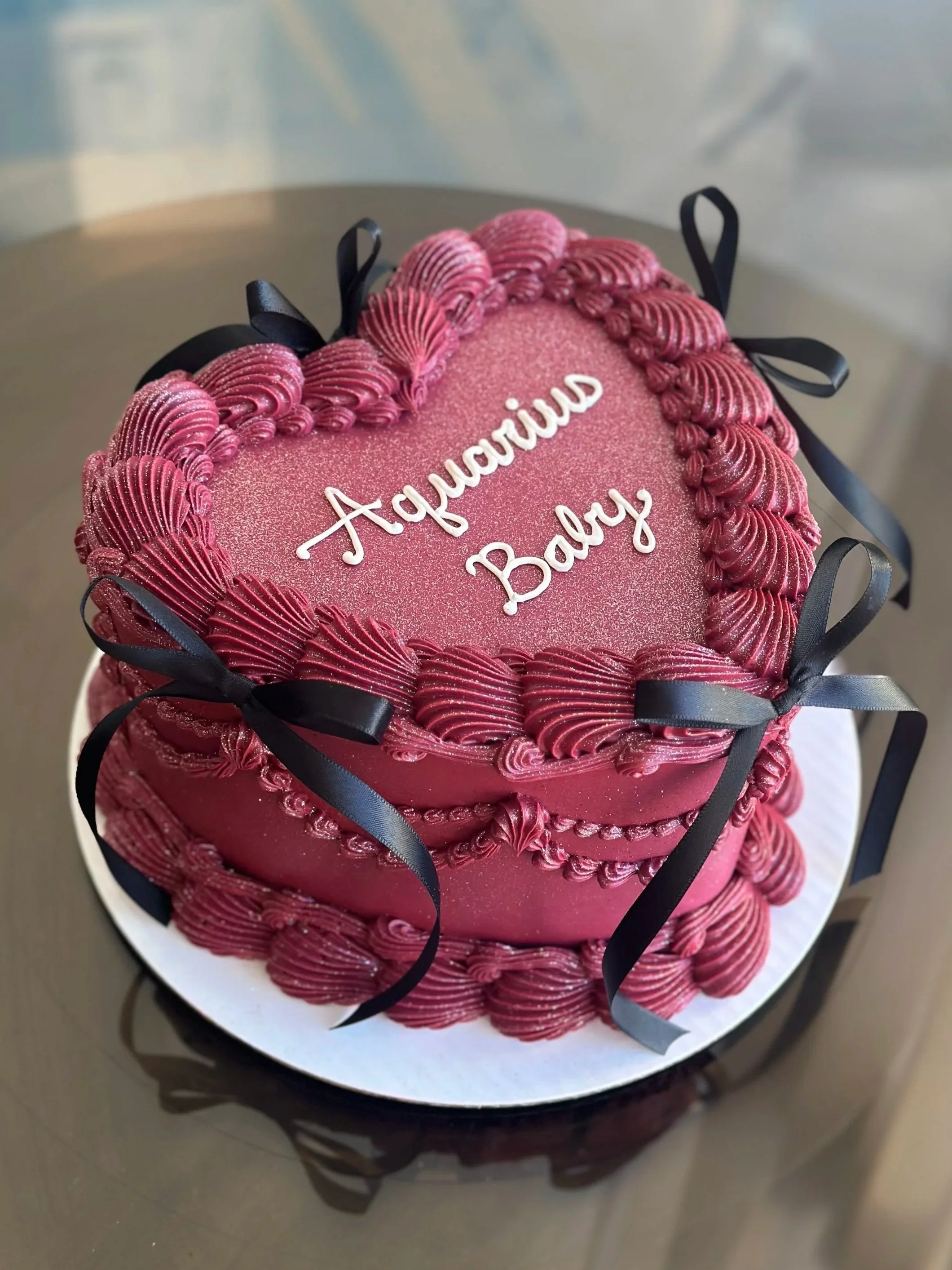 A heart-shaped pink cake decorated with piped icing and black ribbons, with the words 'Auntie Baby' written in white icing on top.