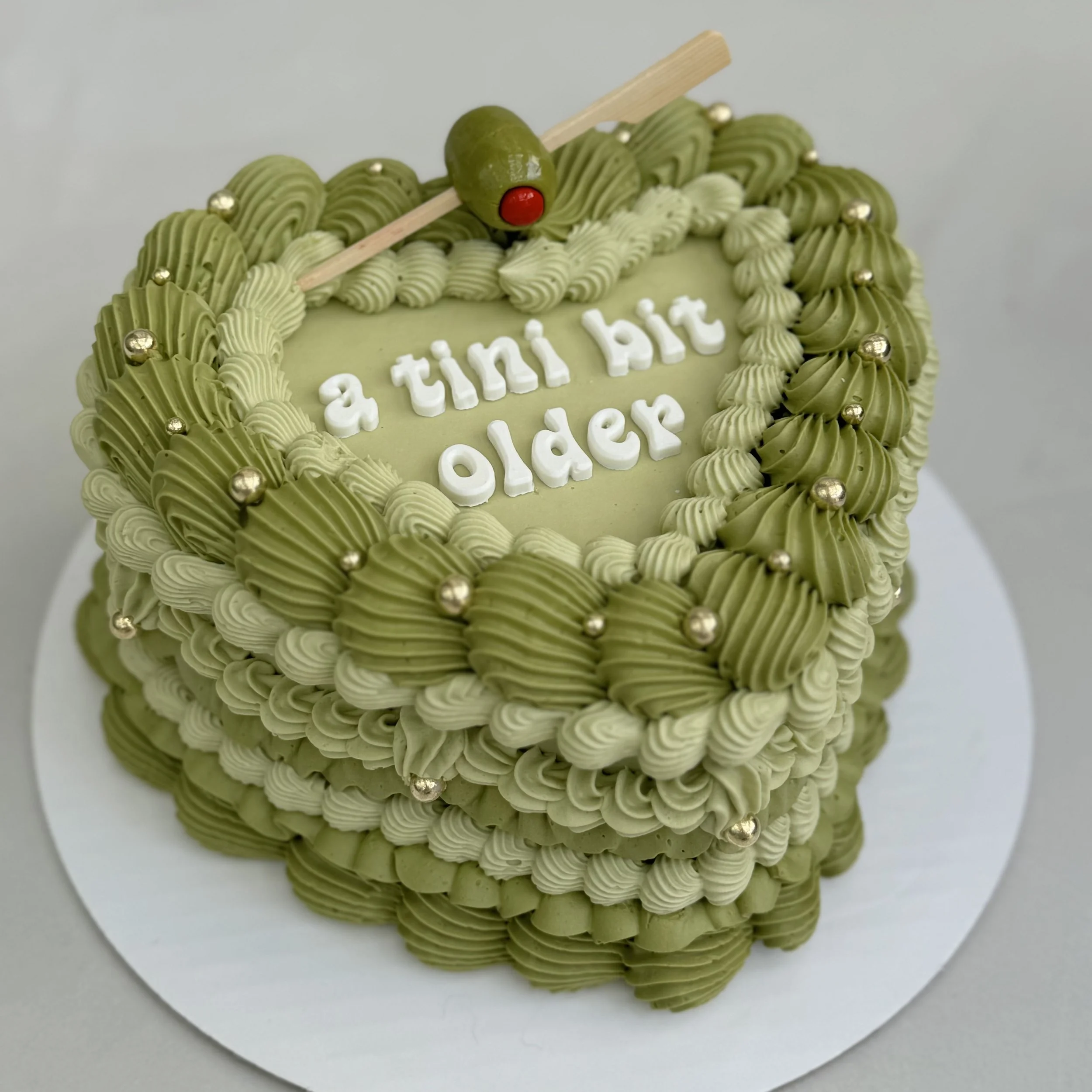 Green birthday cake decorated with piped icing and small silver balls, with a message that reads "a tiny bit older" in white icing and a small olive with a toothpick and cherry on top.